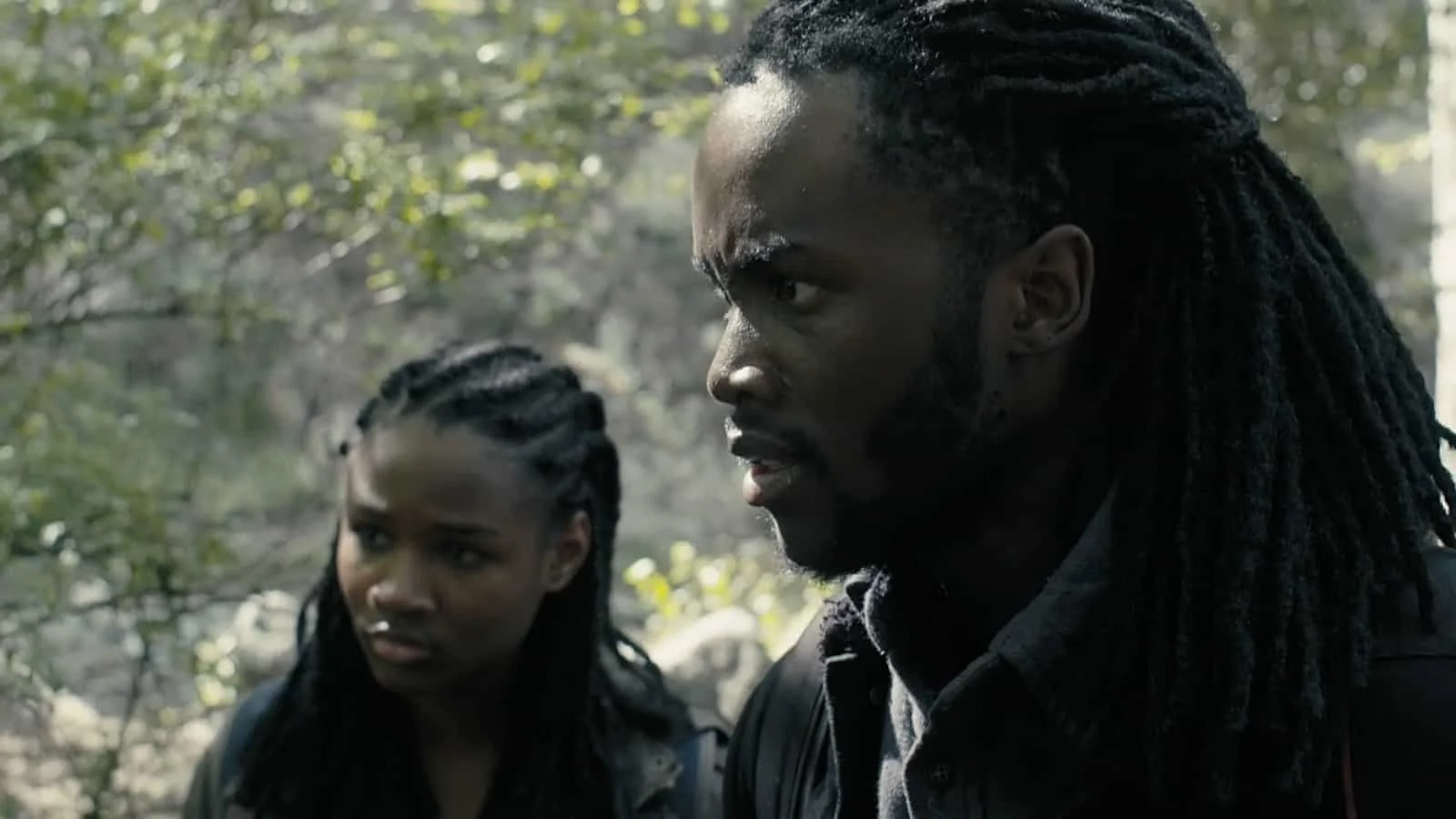 A man and a woman with braided hair are outdoors among trees. The man has long dreadlocks and a serious expression. The woman is looking at him with concern.