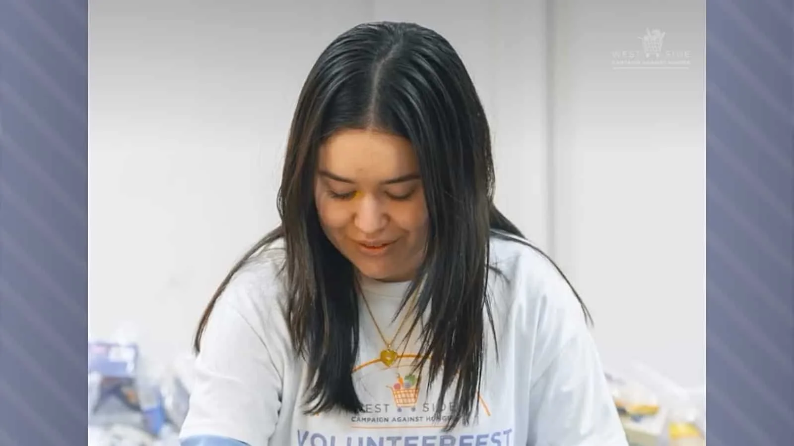 A young woman with long dark hair looking downward, wearing a white t-shirt and a gold necklace, participating in a volunteer event.