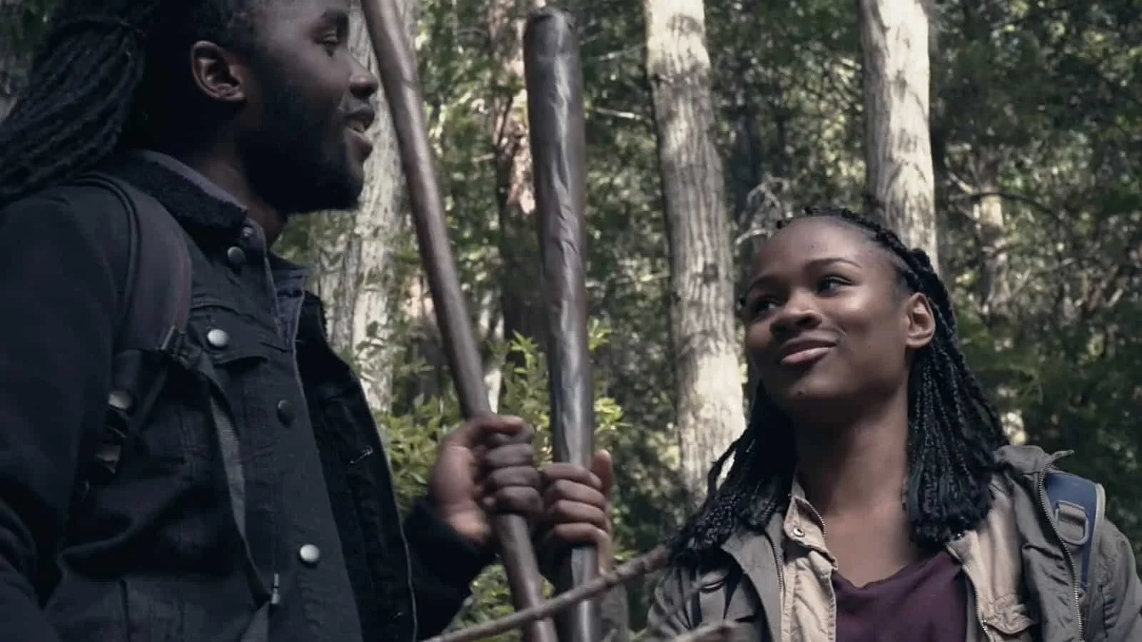 A man and a woman are having a conversation in a forest, with trees in the background. The man has long dreadlocks and is holding a walking stick, while the woman has braided hair and is looking up at him.