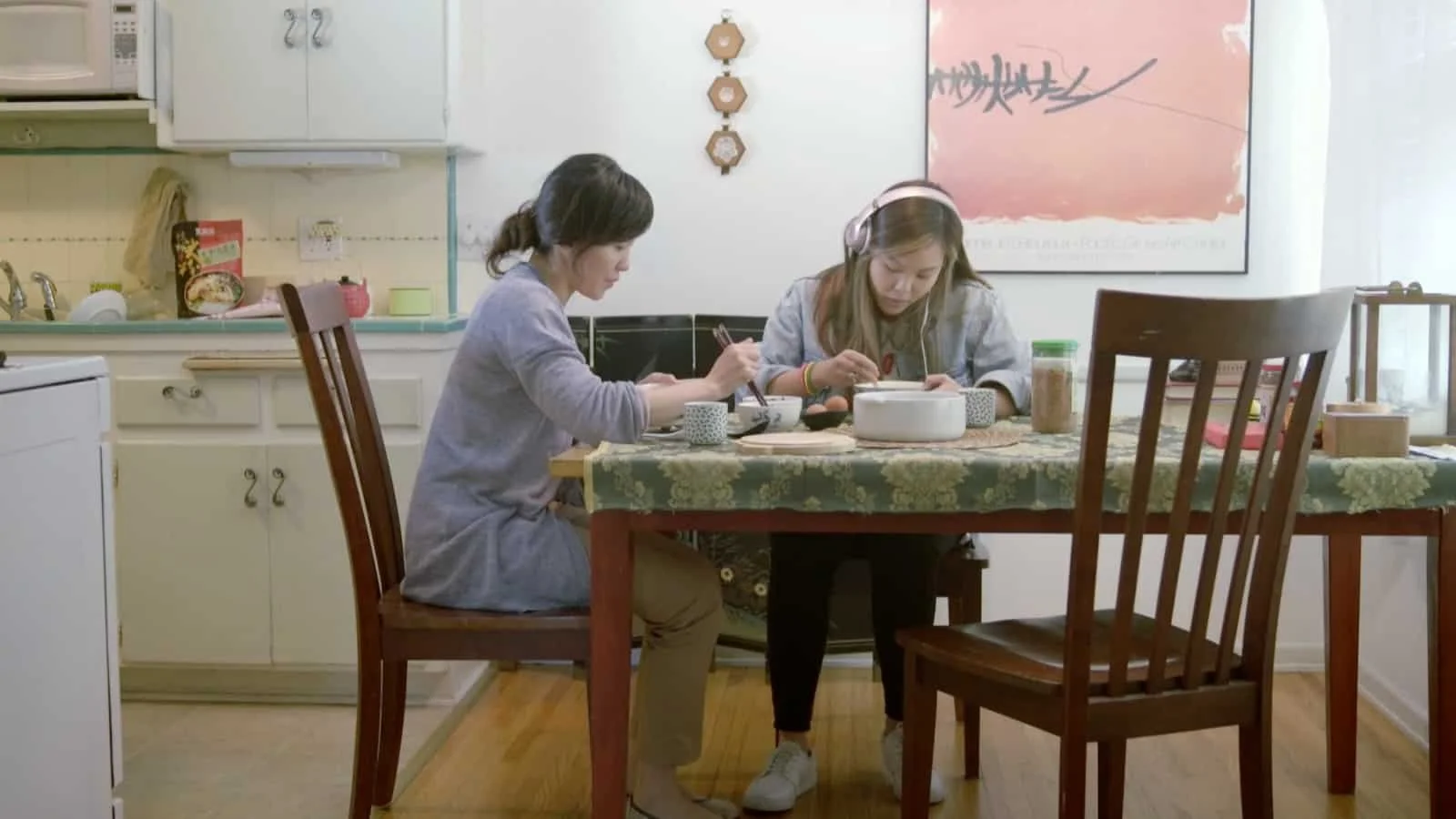 A mother and daughter sit at a wooden dining table eating a meal. They are in a cozy kitchen with light-colored cabinets, a floral tablecloth, and artwork on the wall.
