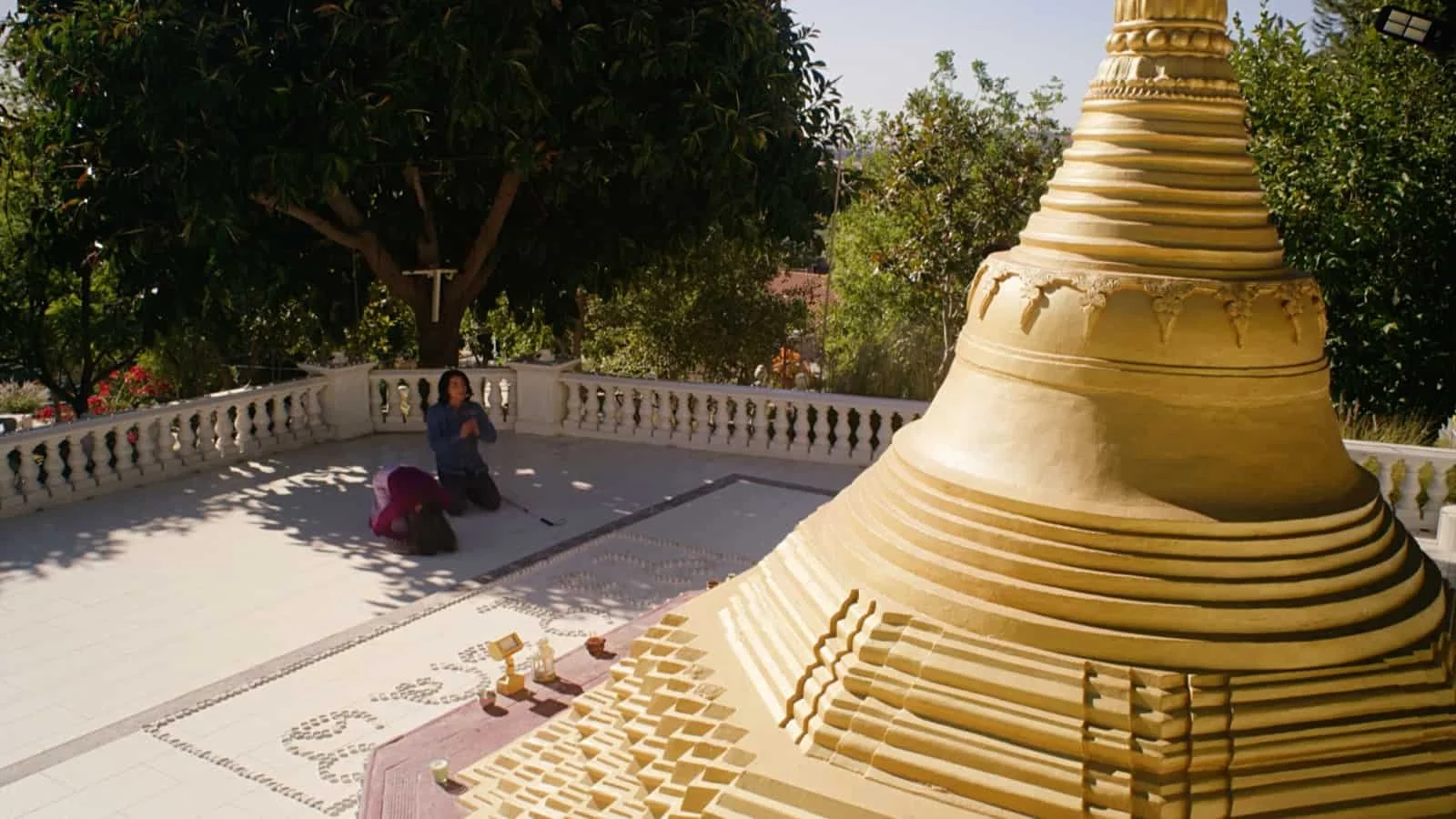 A large, ornate pagoda-style stupa with multiple tiered rings, located outdoors surrounded by trees, with two people kneeling on the ground near the railing.