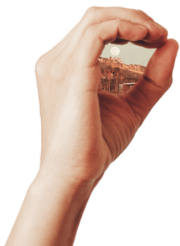 A hand holding a small circle view of a cityscape with houses and trees, with the moon visible in the sky.