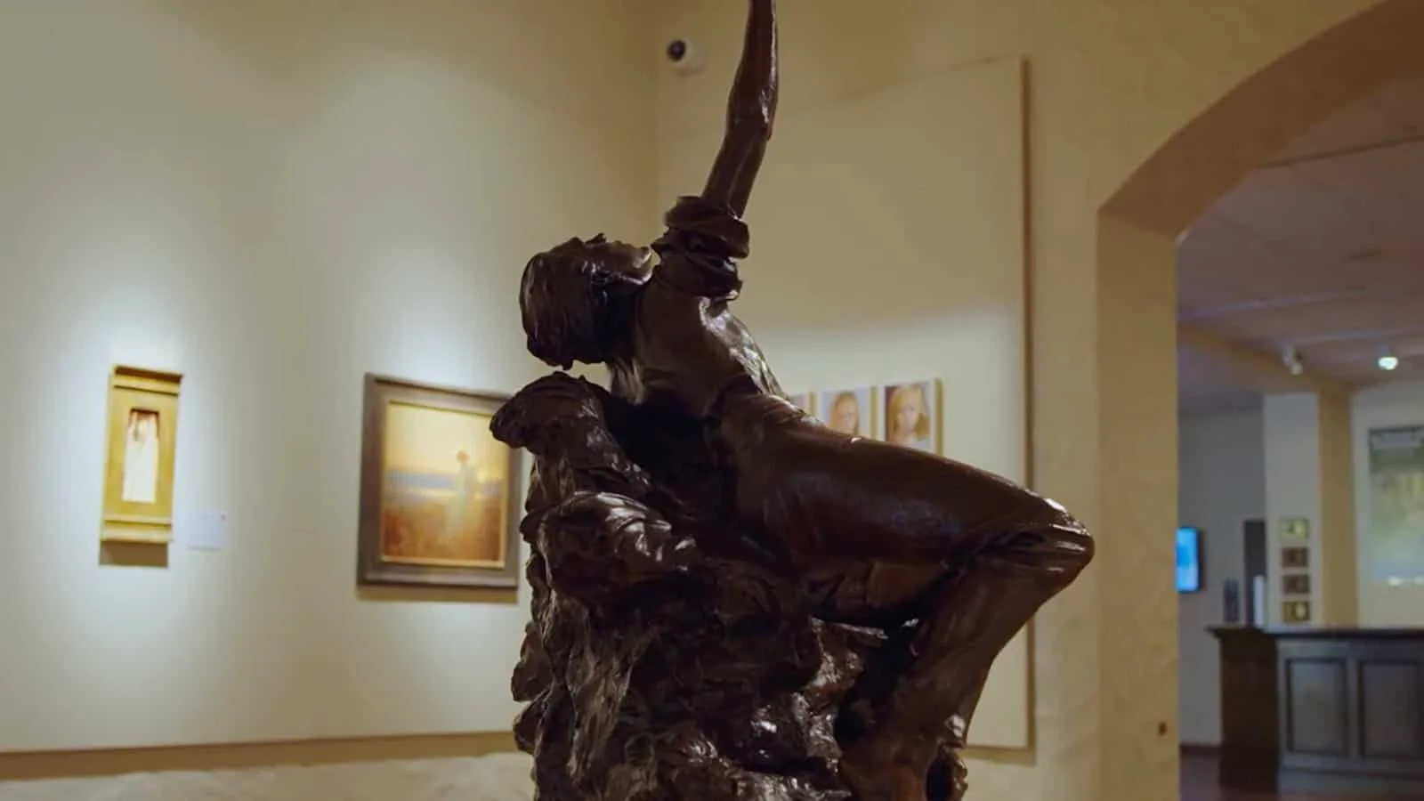 Bronze sculpture of a person in a dramatic pose, with one arm raised and head tilted back, displayed in an art gallery.