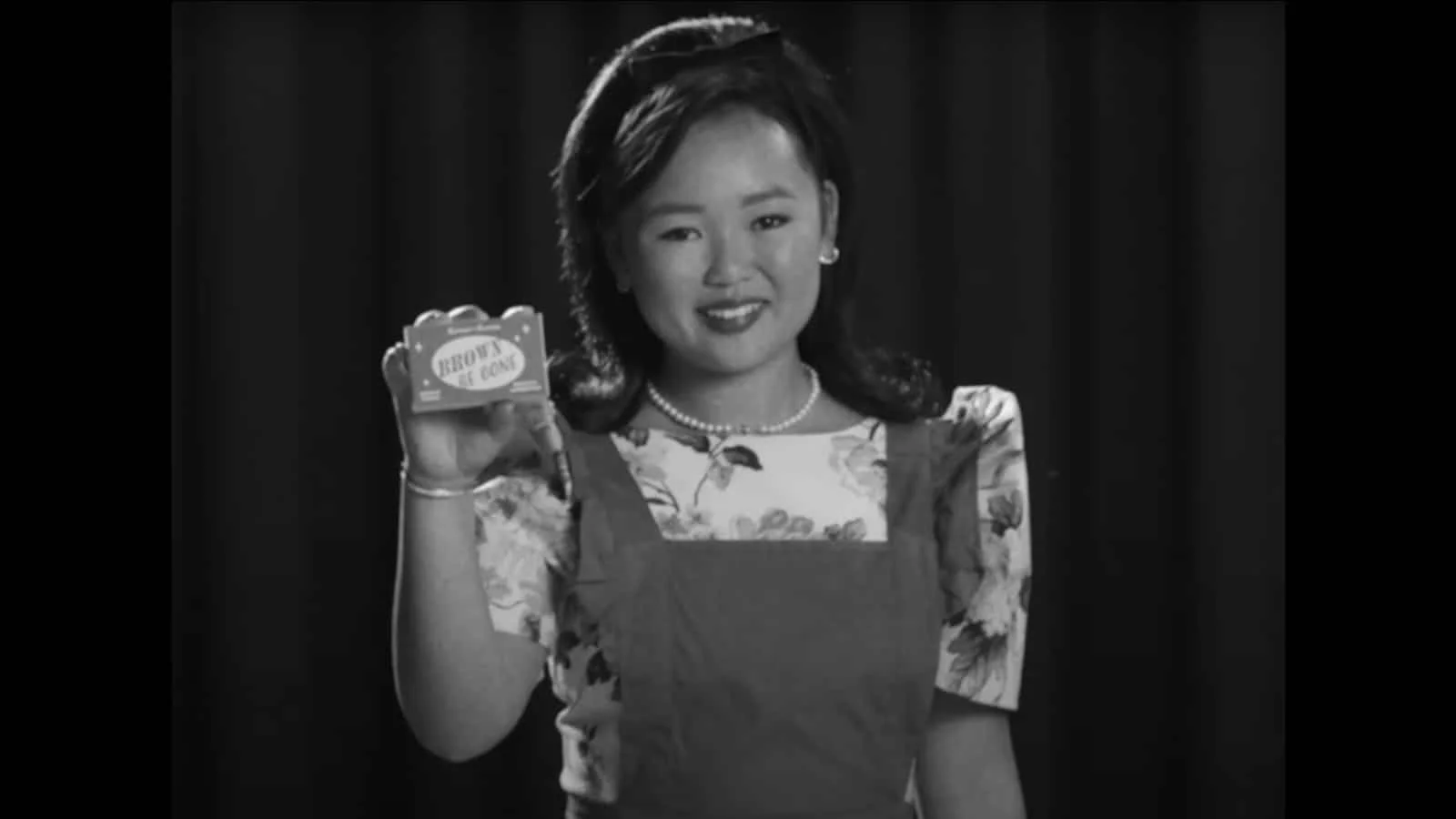 A woman with shoulder-length hair, wearing a floral blouse and a necklace, holding a small that says Brown Be Gone while smiling at the camera, in a black-and-white setting with a dark background.