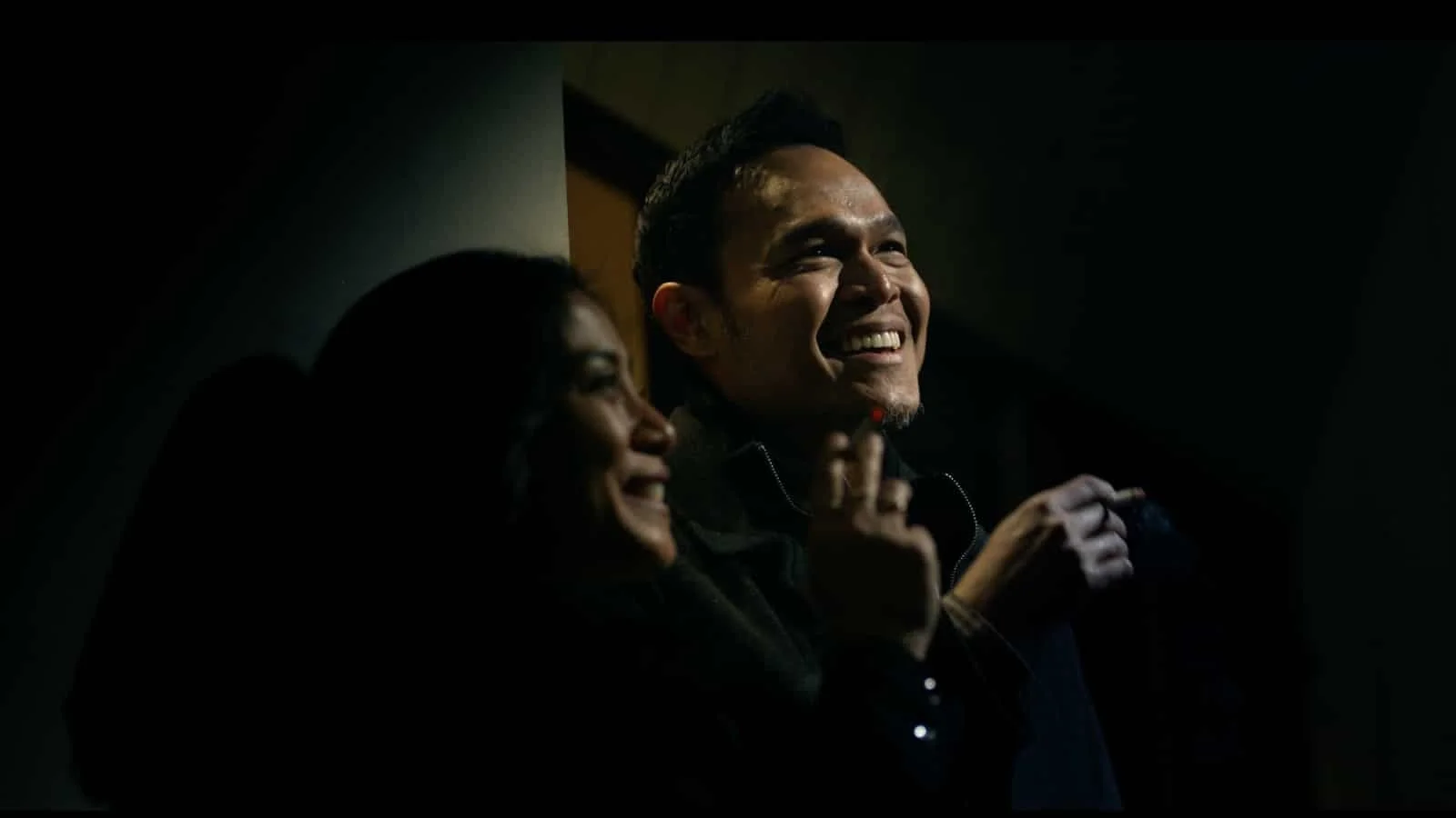 Two smiling people, a man and a woman, in a dark room. The man is facing forward and the woman is partially visible, both appear happy and engaged.