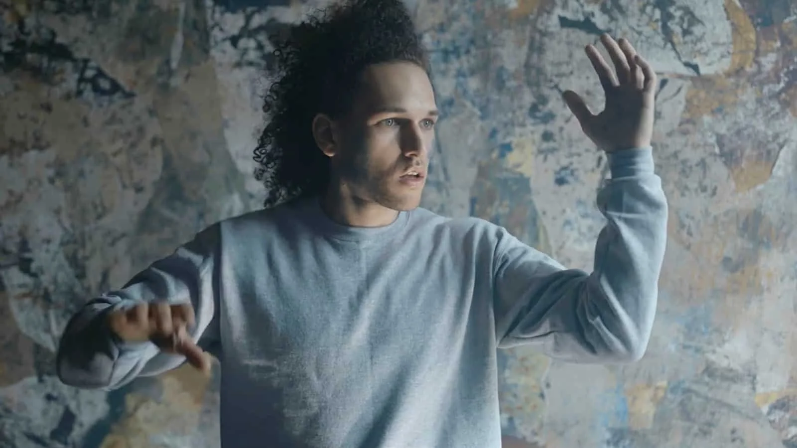 Young man with curly hair raises his right hand, standing in front of a textured wall.