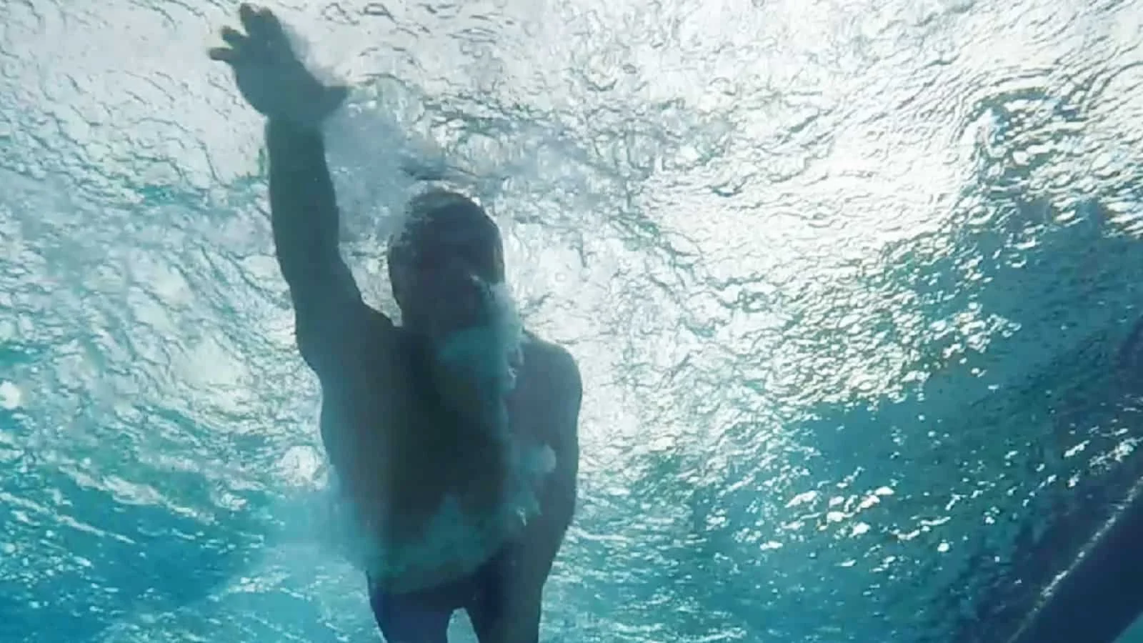 A person underwater, reaching forward with one arm, with water surface above and light filtering through.