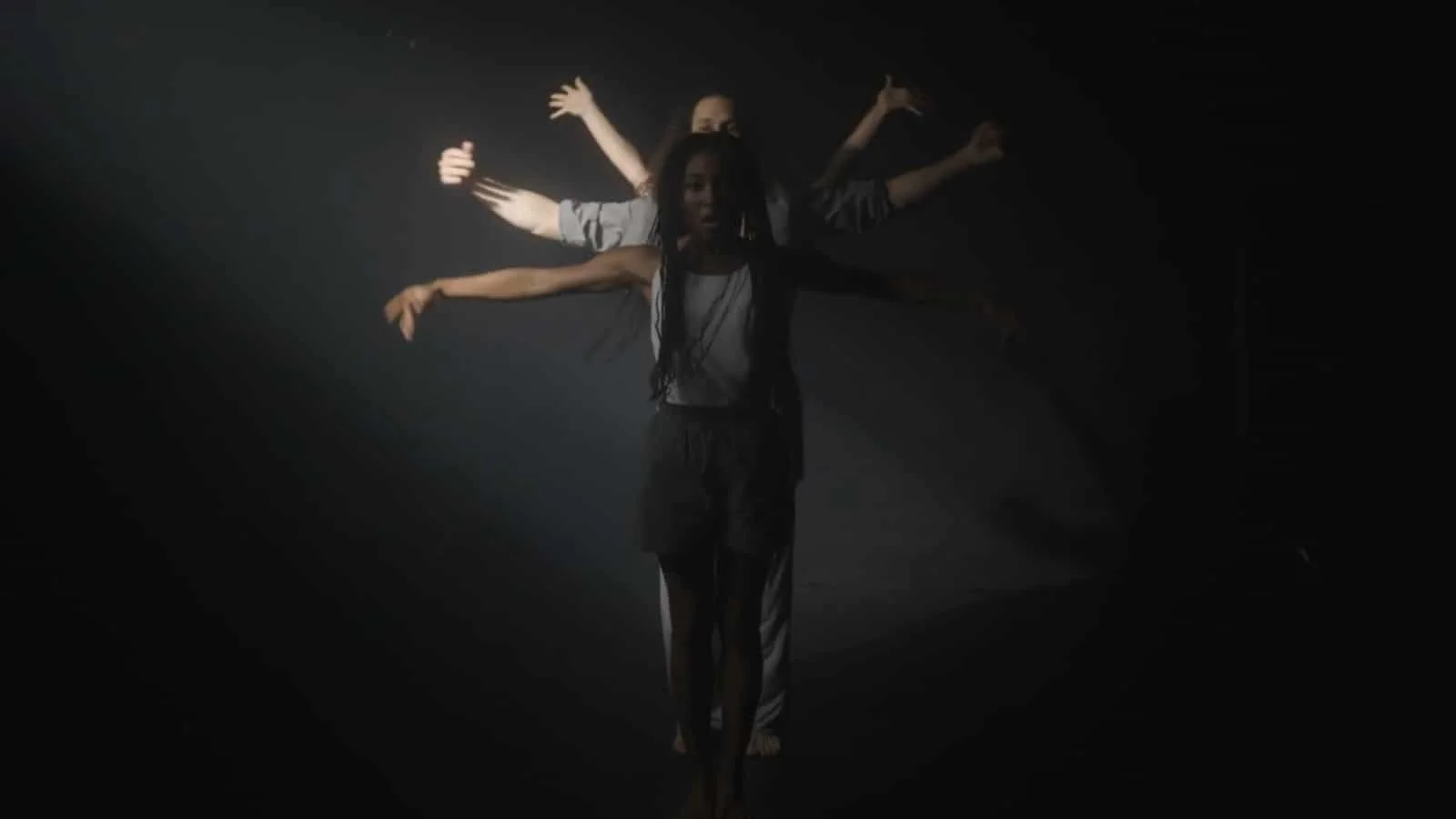 A woman standing with her arms outstretched in front of a dark background, with multiple arms of other people extending from behind her, creating a visual effect similar to a multi-armed figure.