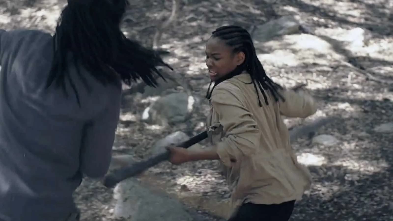 A young women and a man in dreadlocks fighting outdoors on rocky ground, one holding a stick, with expressions of distress or anger.