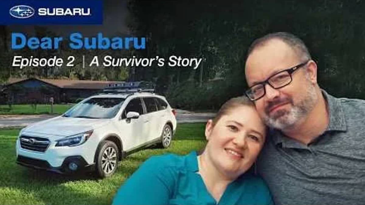 A woman and a man smiling and hugging in front of a white Subaru SUV parked on grass with trees and a building in the background, with text that reads "Dear Subaru, Episode 2 | A Survivor's Story."