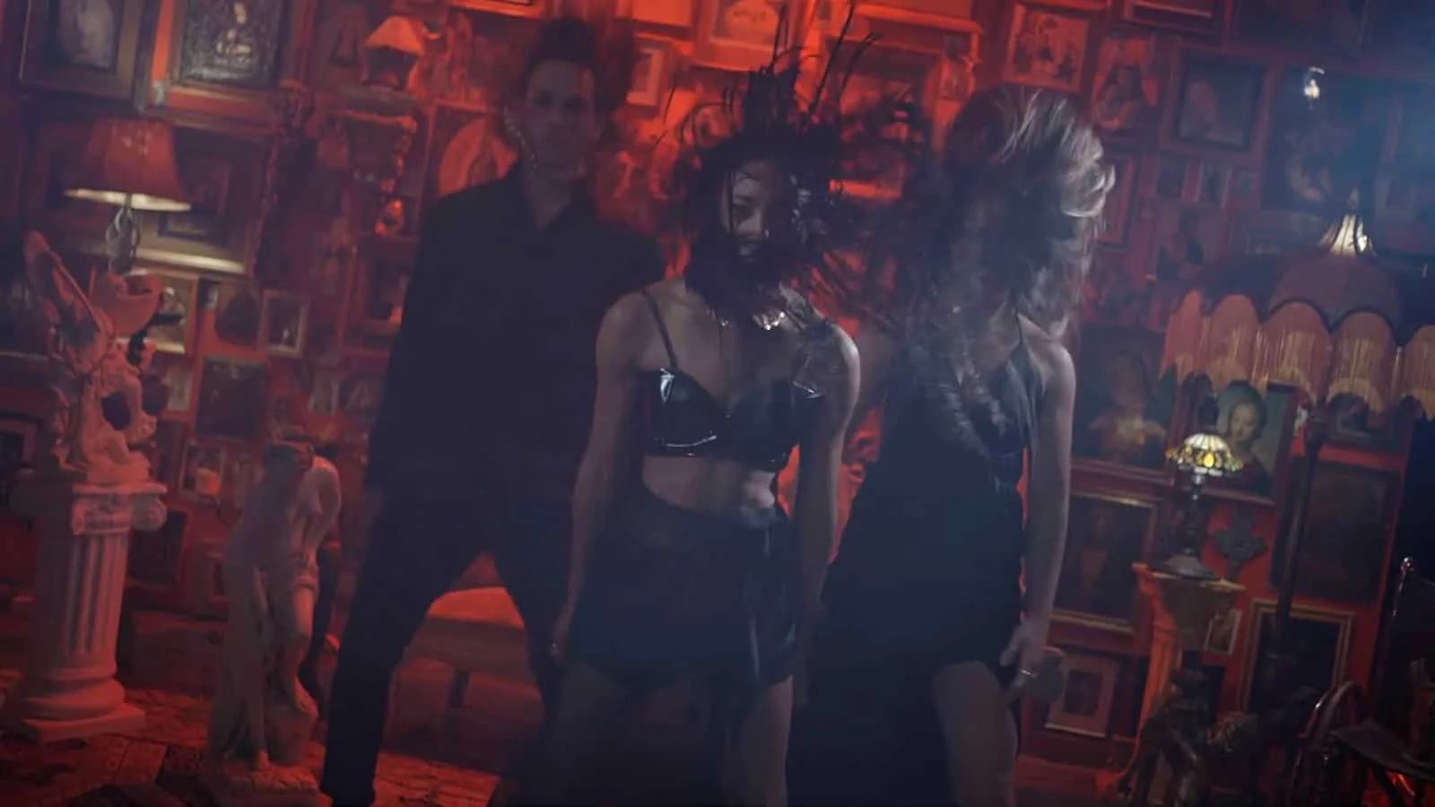 Two women and a man dancing in a dimly lit room with red and orange lighting, decorated with numerous framed pictures, statues, and vintage lamps on the walls and furniture.