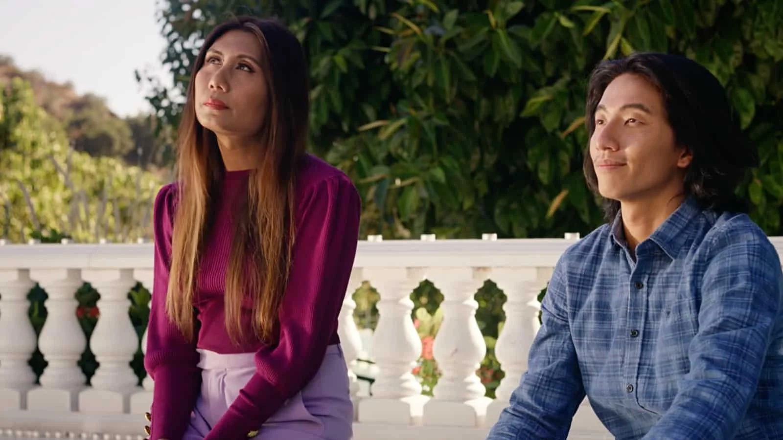 A woman and a man sitting outdoors near a white railing with green trees in the background.