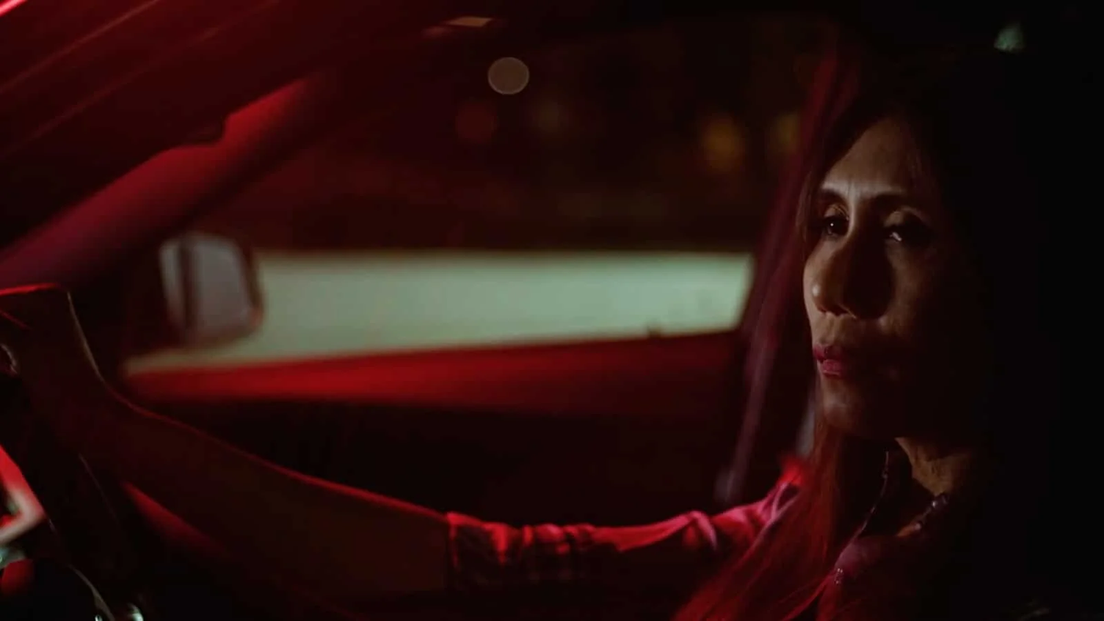 A woman with red hair driving a car at night, illuminated softly by the interior and exterior lights.