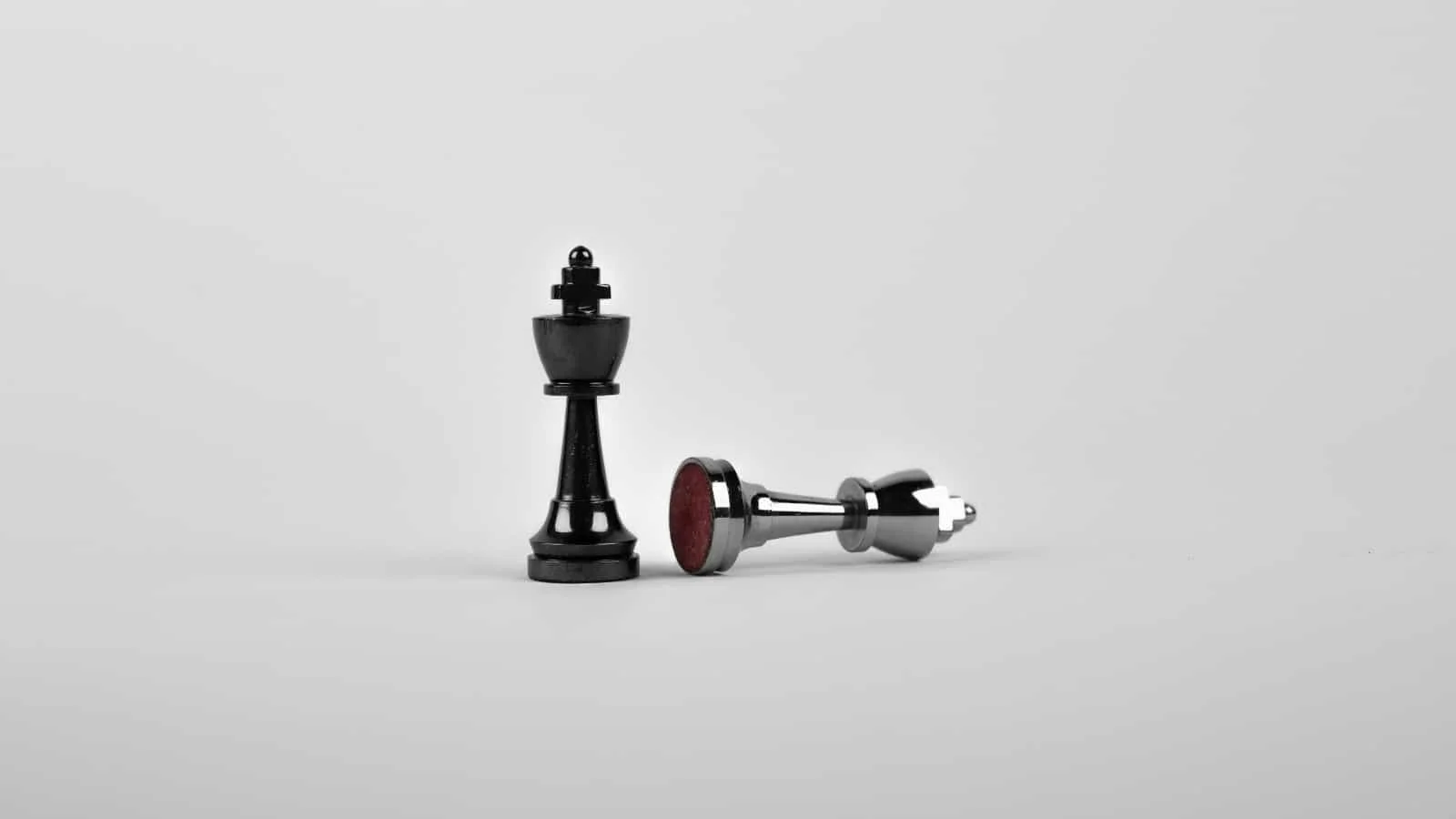 Two black chess pieces, a black king standing upright and a fallen chrome king, on a plain light gray surface with a neutral gray background.