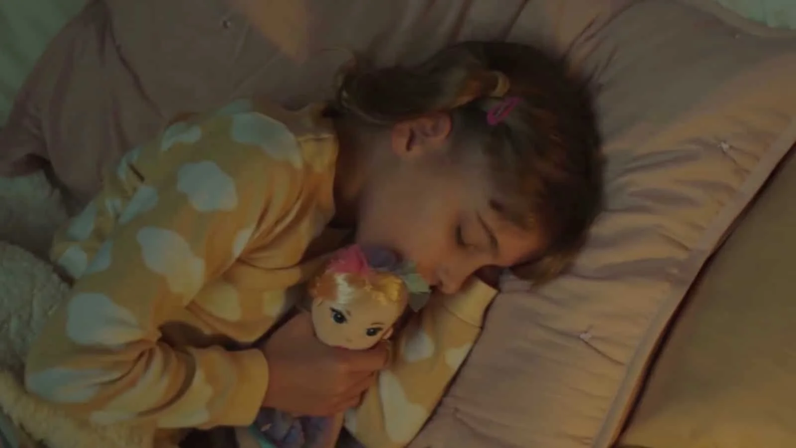 A young girl sleeping on a bed, hugging a doll with orange hair, wearing a yellow pajama with white and pink patterns, with a pink hair tie and long hair.