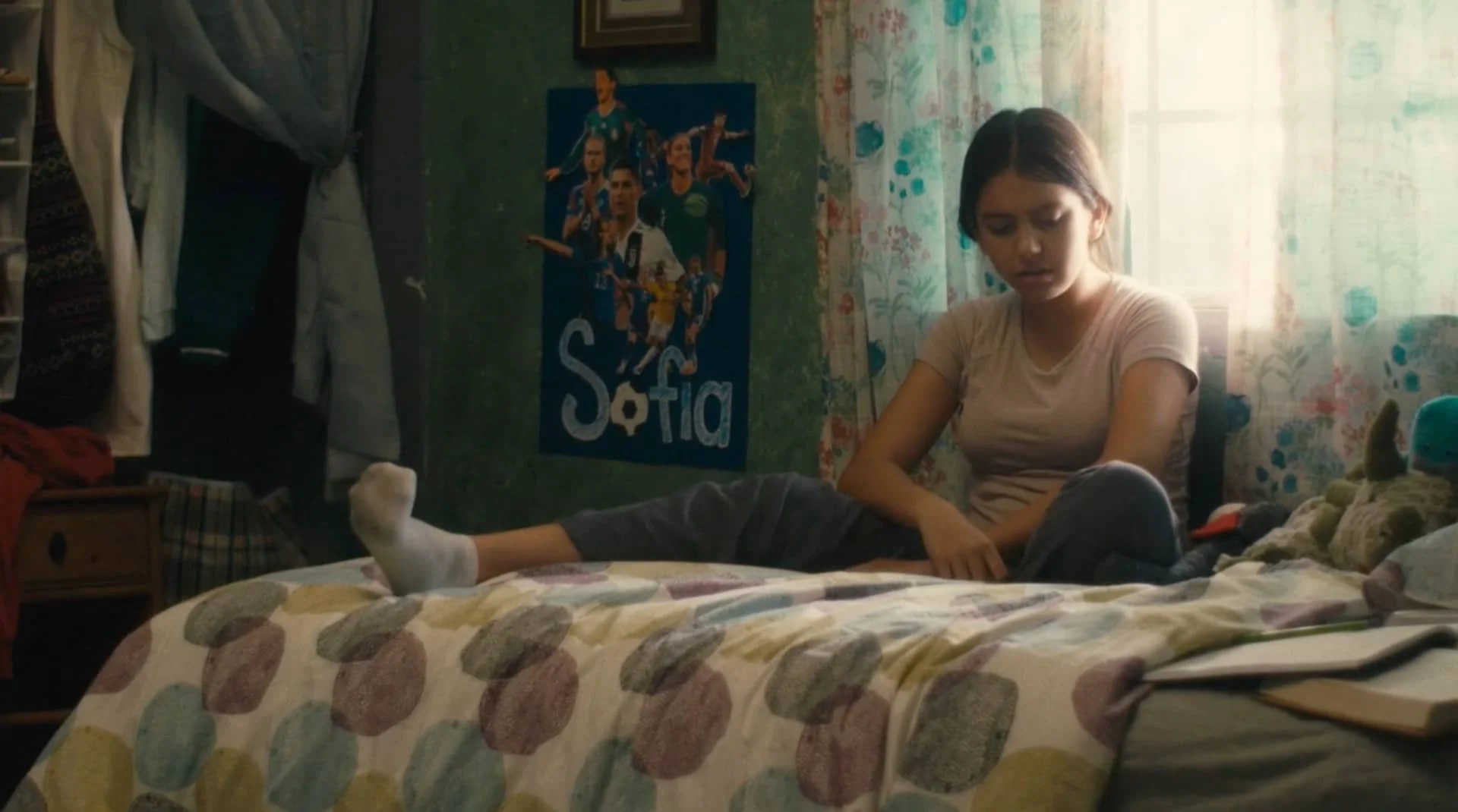 Teenage girl sitting on bed with a colorful quilt, looking downward, in a bedroom with floral curtains, a poster, and closet with hanging clothes.