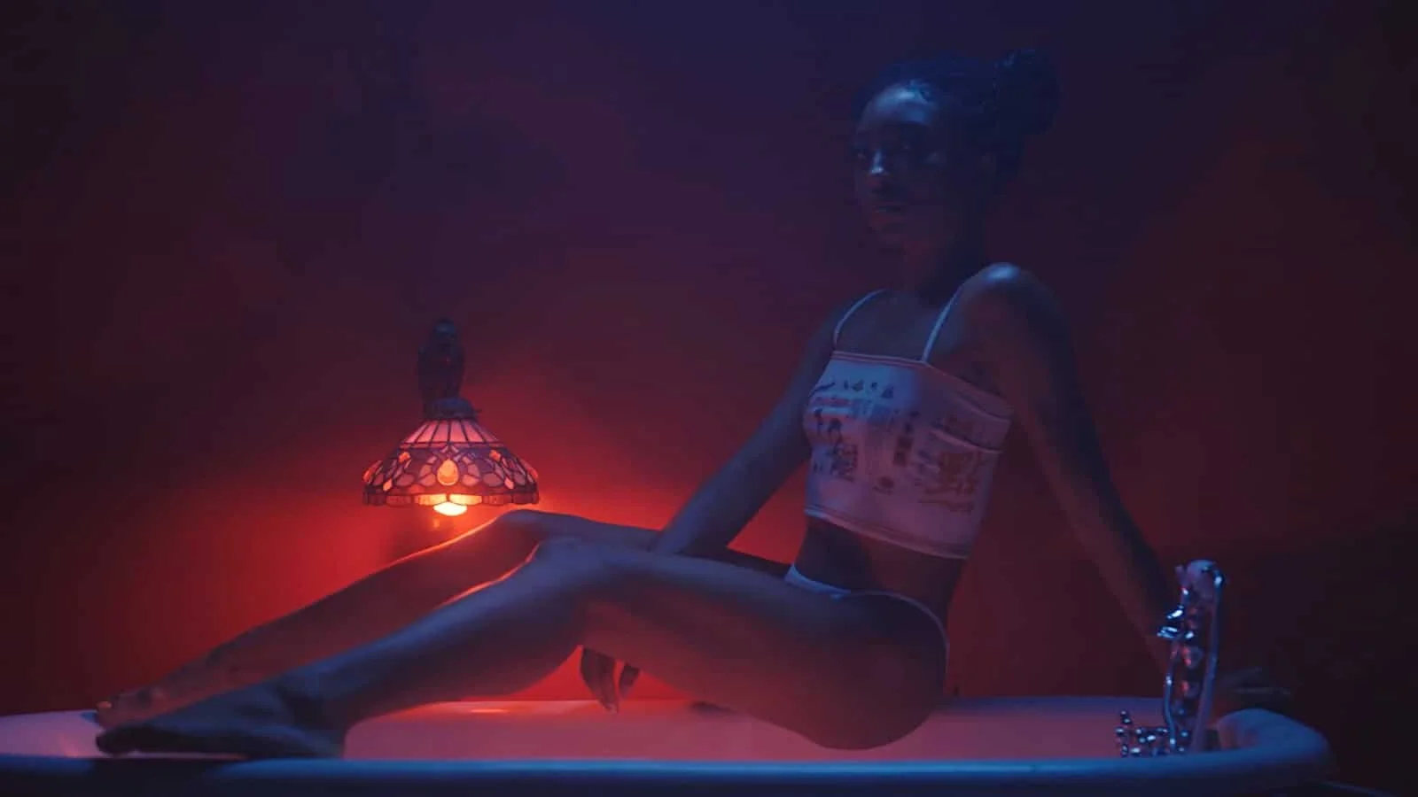 A woman sitting on the edge of a bathtub under dim red and purple lighting, with a small lamp nearby.