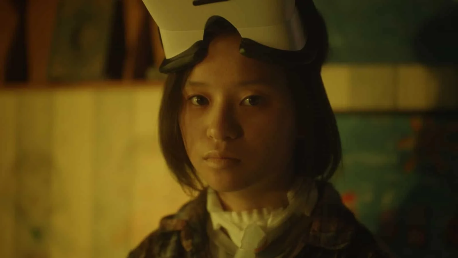 A woman with short dark hair looking directly at the camera, wearing a VR headset, in a dimly lit room with yellow and green tones.