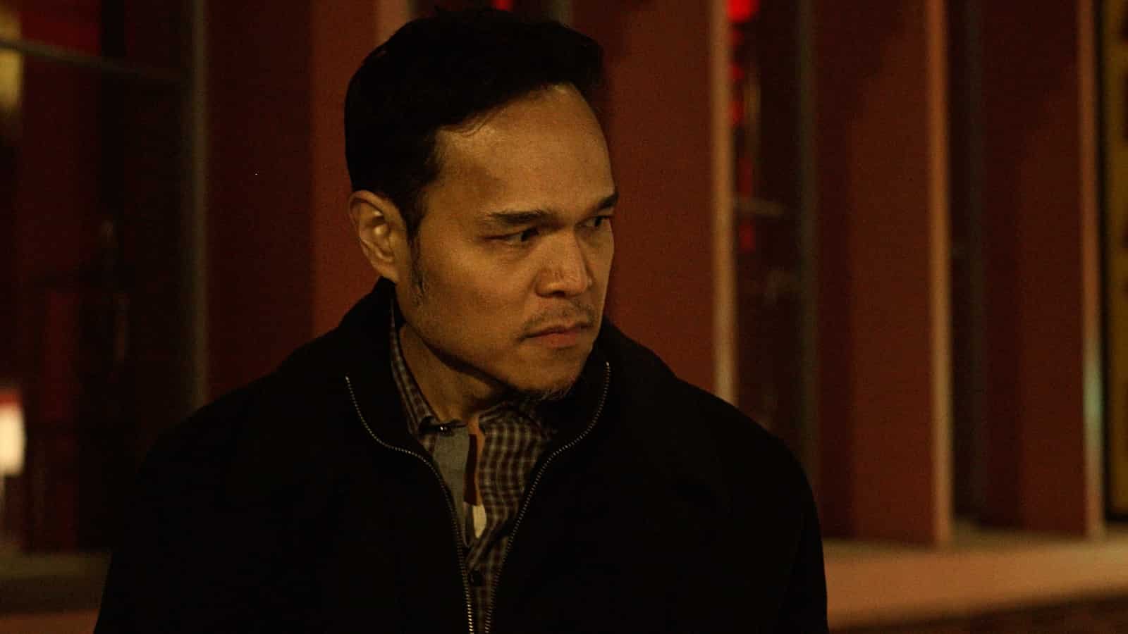 A man with dark hair and a light beard looking to the side, wearing a dark jacket over a plaid shirt in a dimly lit room.