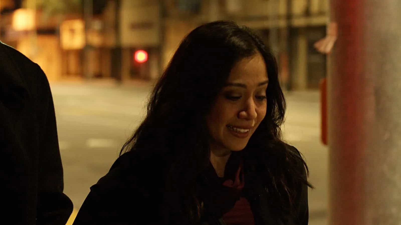 A woman with long dark hair, smiling and looking down, standing outside at night near a pole with blurred background of a building with lights.