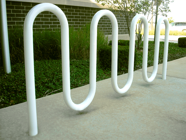 White metal bike rack on a concrete sidewalk outdoors with grass and trees in the background.