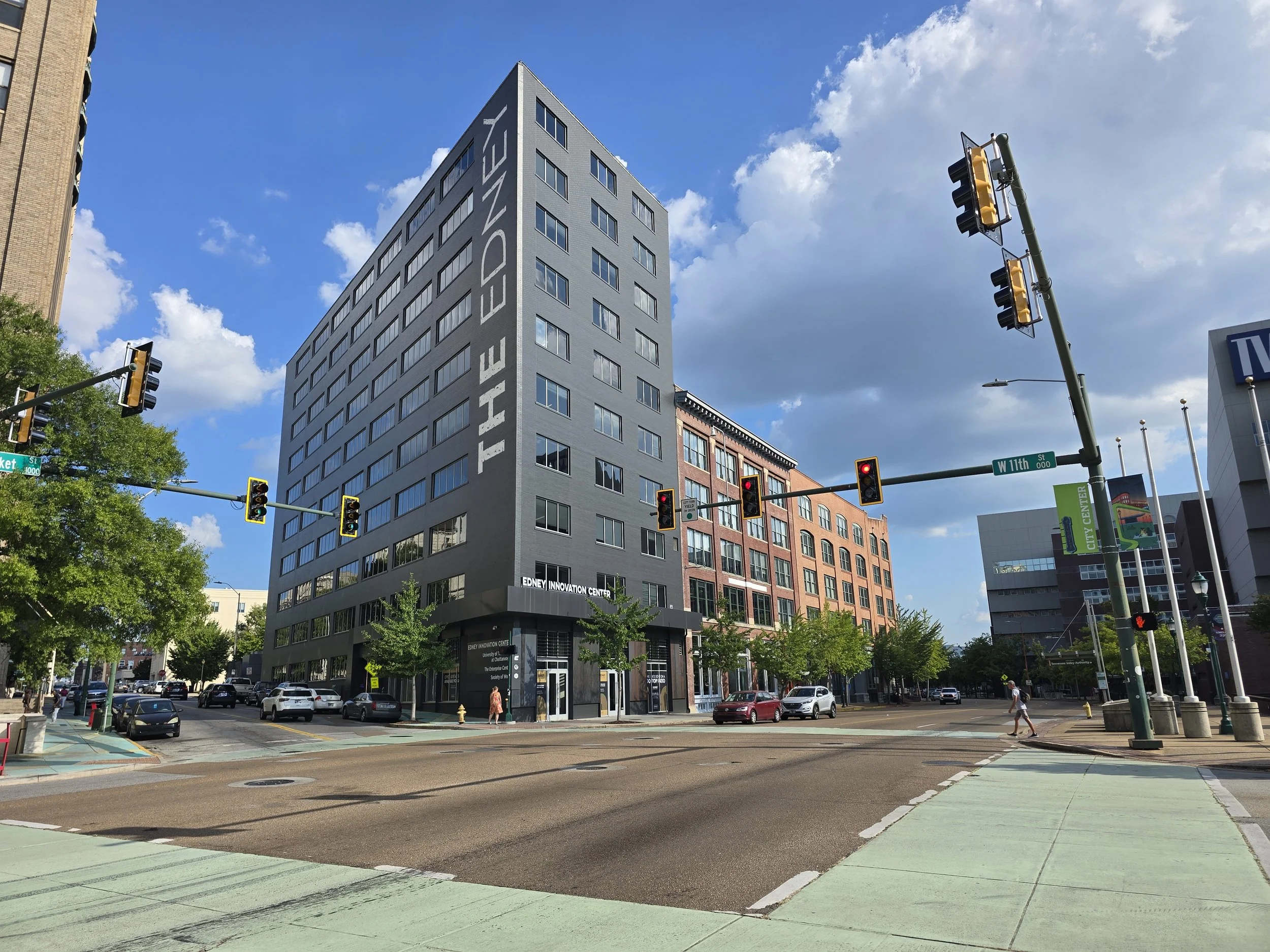 City street intersection with traffic lights, people crossing, cars, and modern buildings, including Edney Innovation Center with blue sky and clouds