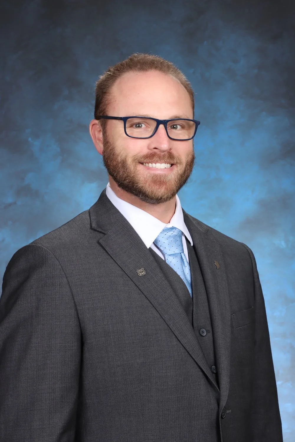 A man in a gray suit, light blue shirt, and light blue tie, wearing glasses, smiling, against a blue textured background.