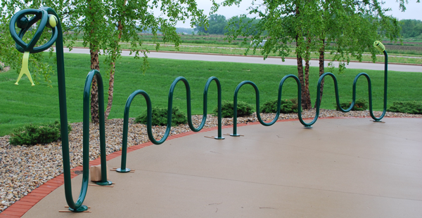 Outdoor bike rack with a wavy design, installed on a concrete sidewalk, with trees and grass in the background.