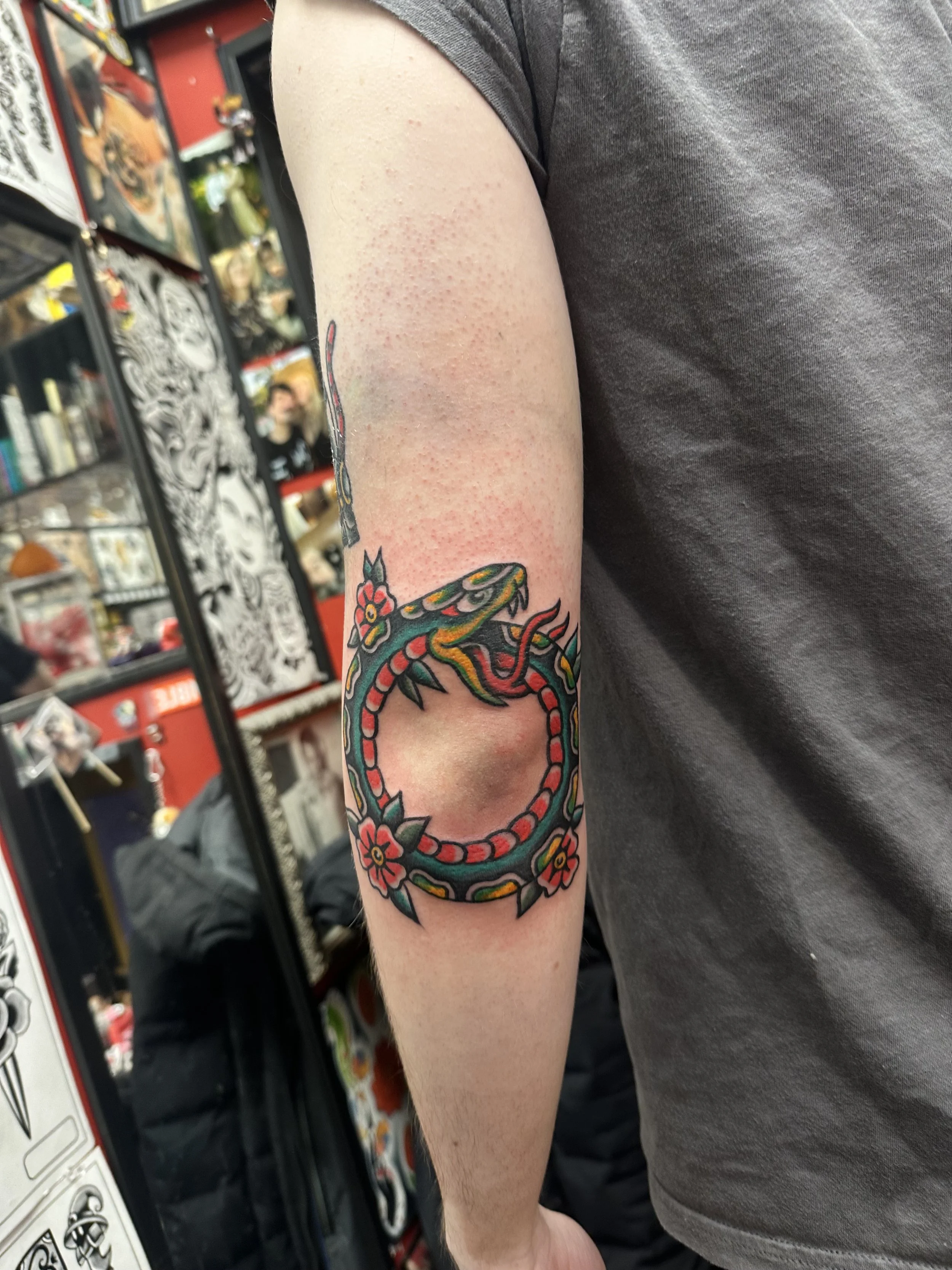 A person's upper arm with a colorful tattoo of a snake intertwined with flowers. The background shows blurred shelves with various items, indicating the setting is a tattoo shop or similar space.