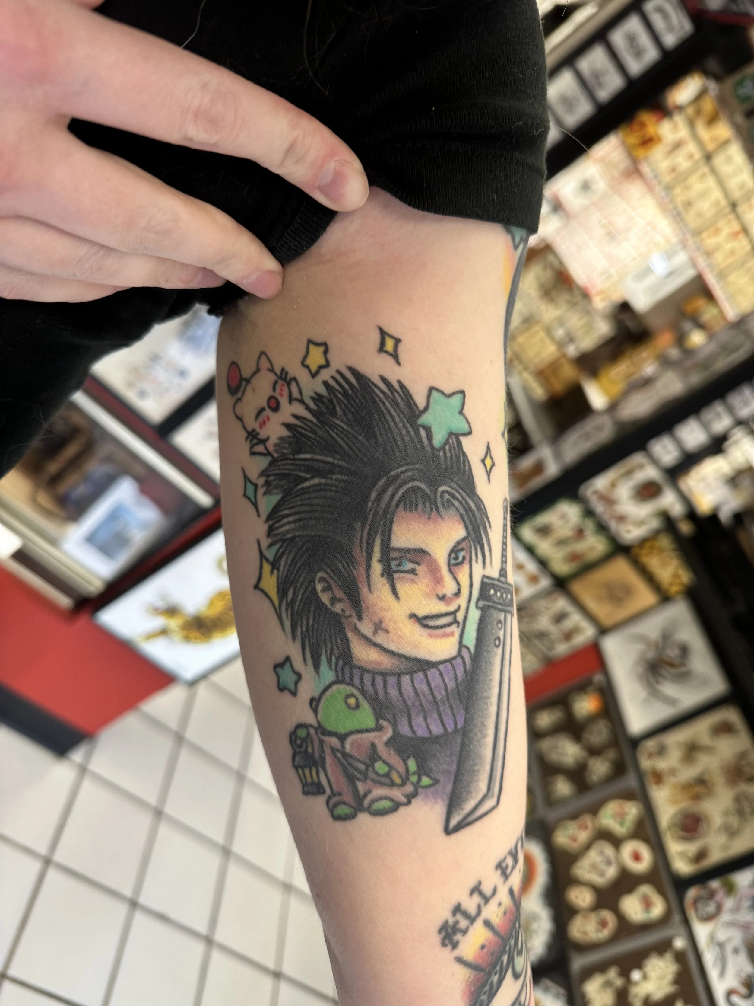 A person's arm with a colorful tattoo of a woman with spiky hair, surrounded by stars and cartoon characters including a mouse, a green creature, and a knife, with a store shelf in the background.