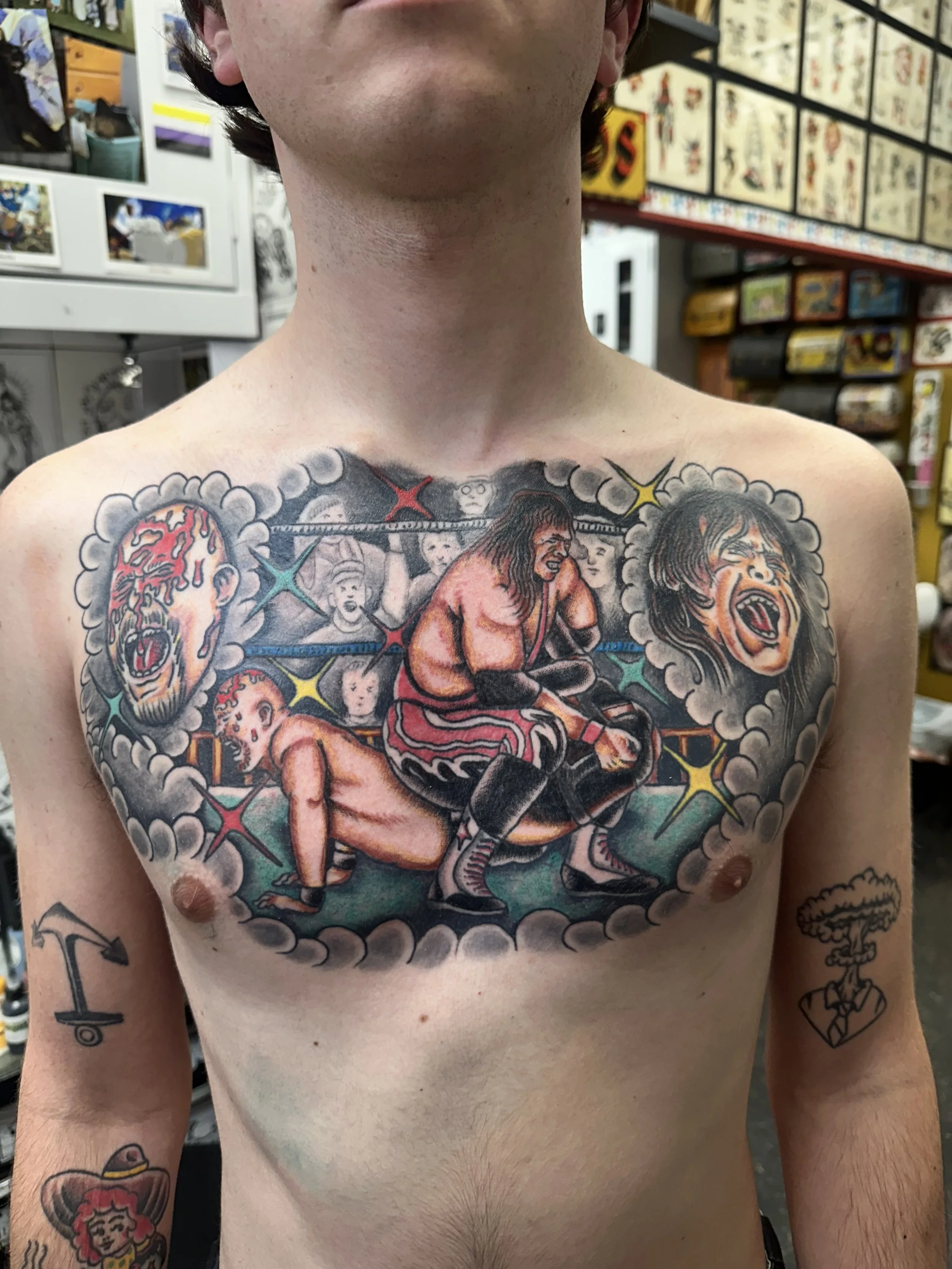 A shirtless man with tattoos on his chest and arms stands in a room filled with colorful items and artwork. The tattoo on his chest depicts wrestlers in a wrestling ring. His left arm has tattoos including a woman with a hat and a cloud with a lightn