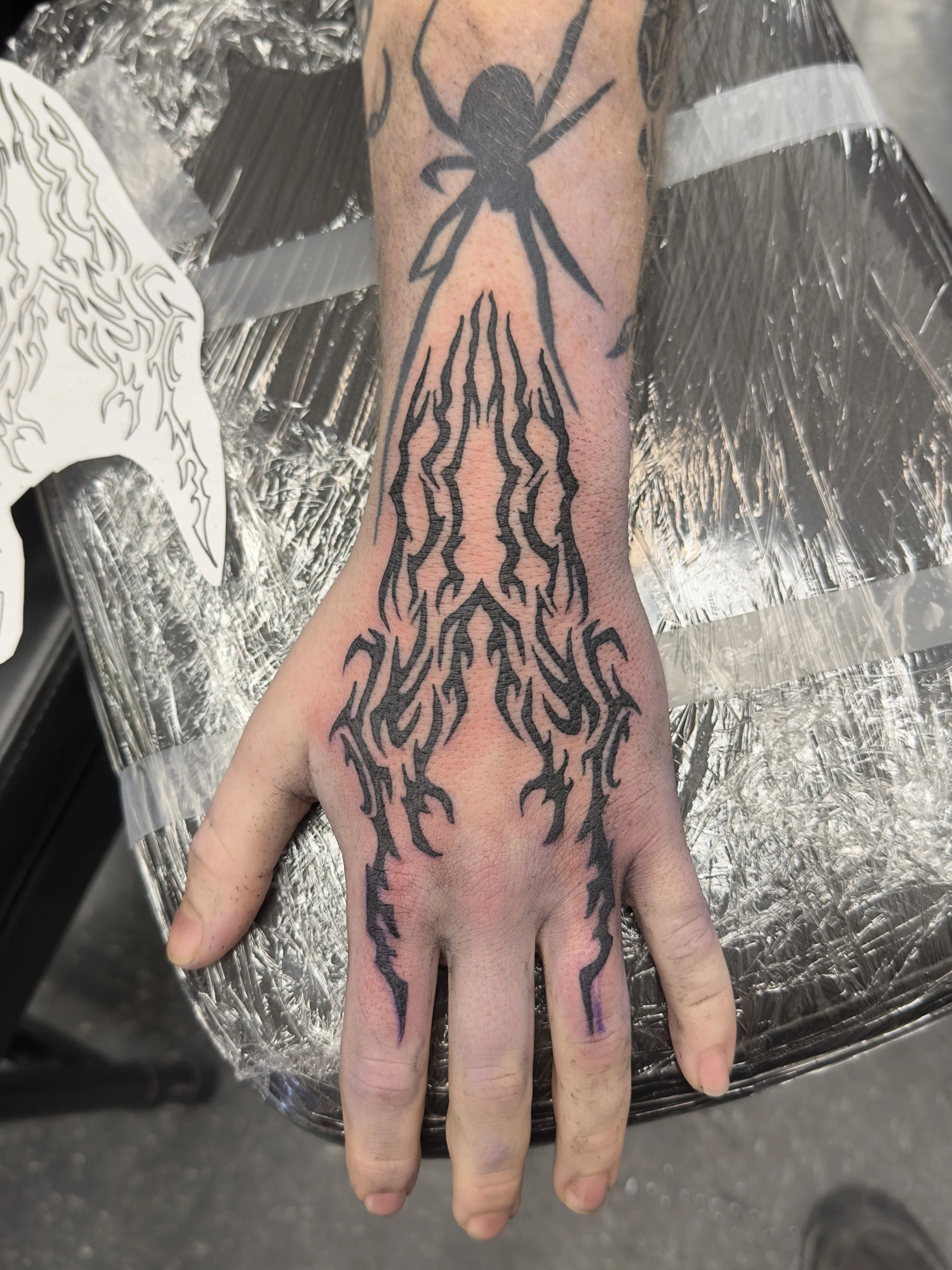 A person's hand with a large black tattoo of a spider on the wrist, extending down to the fingers, with a design resembling claws or lightning bolts.