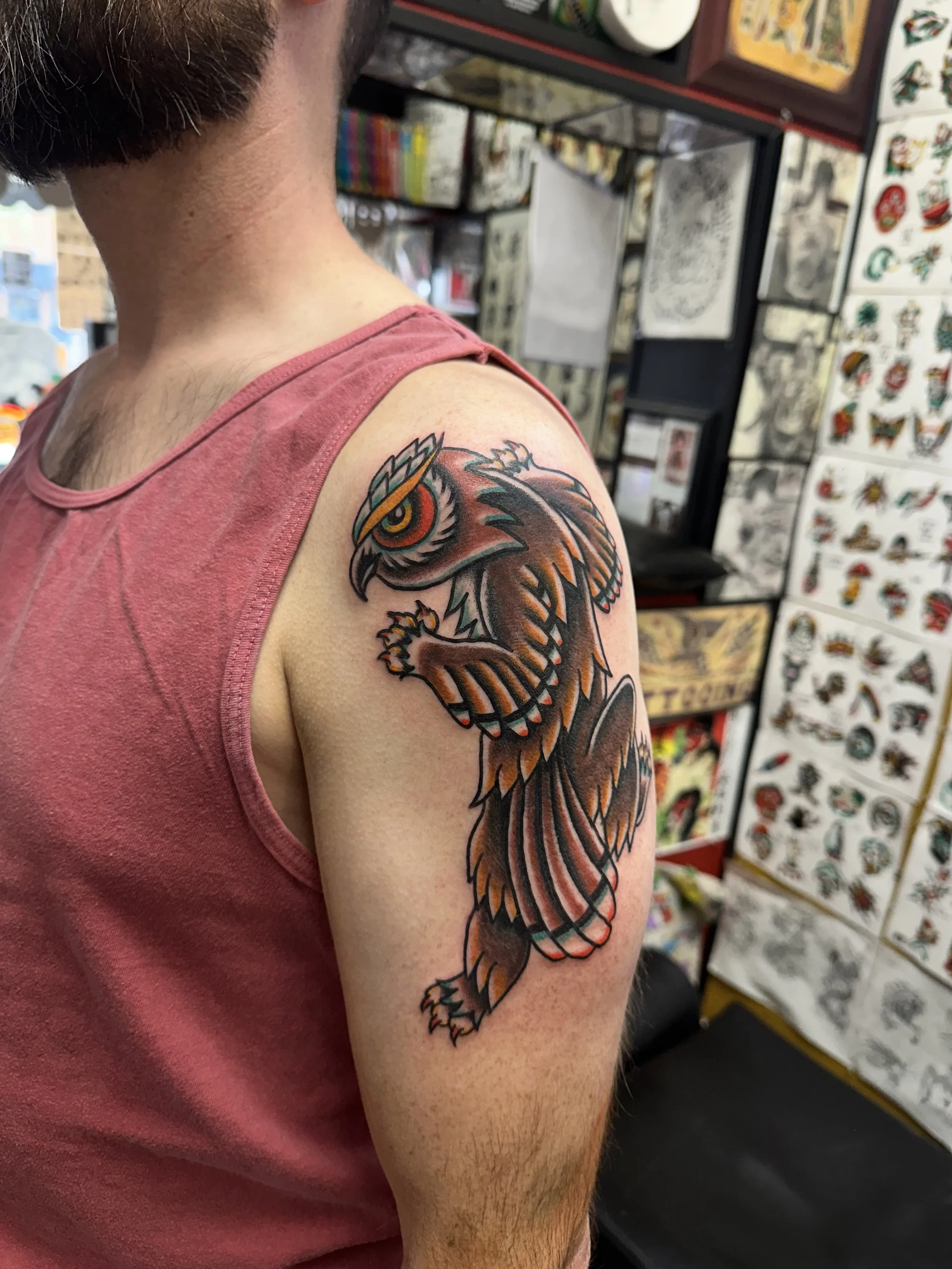 A person with short dark hair and a red tank top showing a colorful tiger tattoo on their upper arm. The background features tattoo flash art and prints displayed on the wall.
