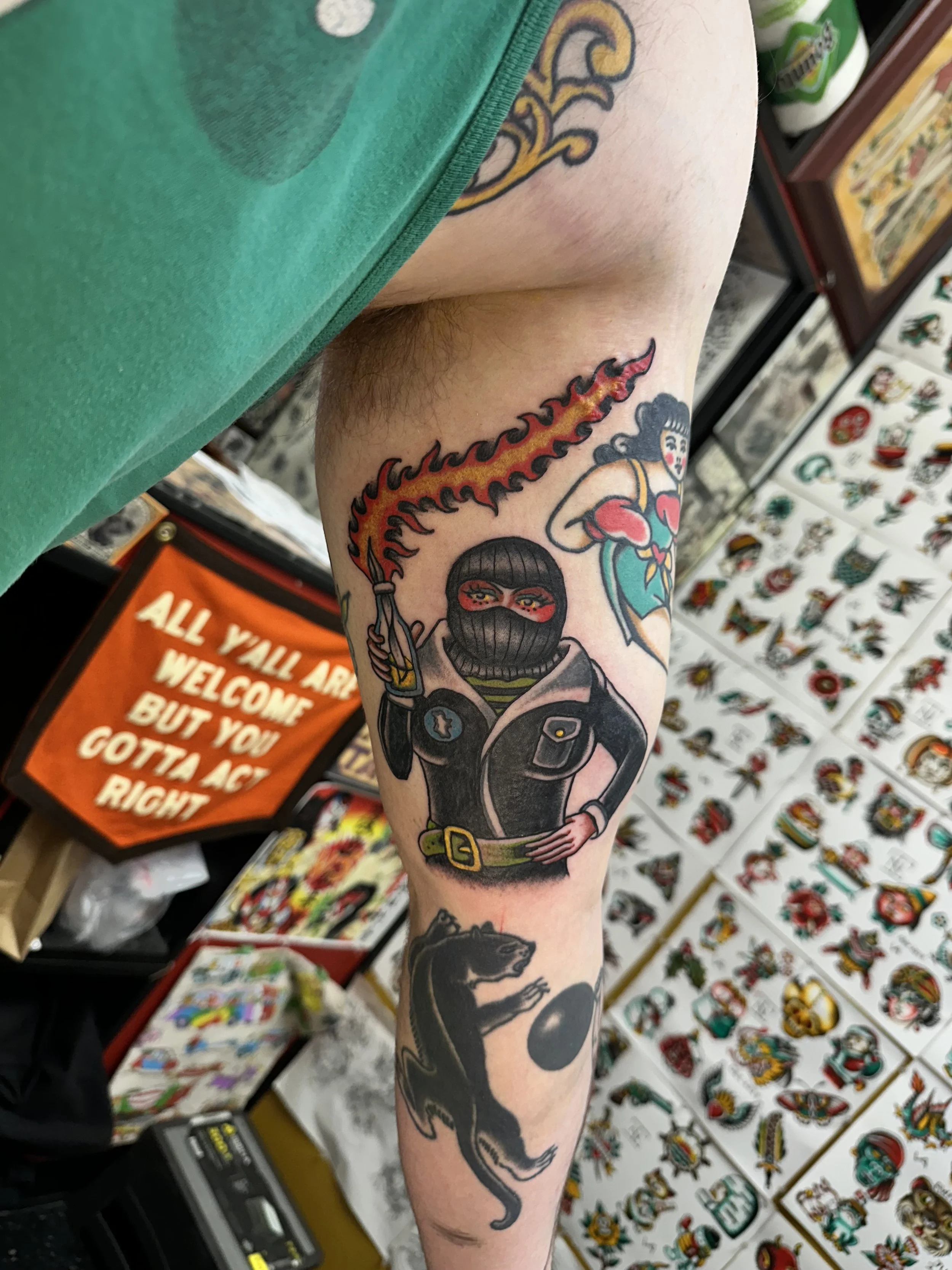 Tattooed arm with a colorful tattoo of a ninja woman holding a sword, a dragon with flames, and a black panther. Background includes tattoo art displays on walls.