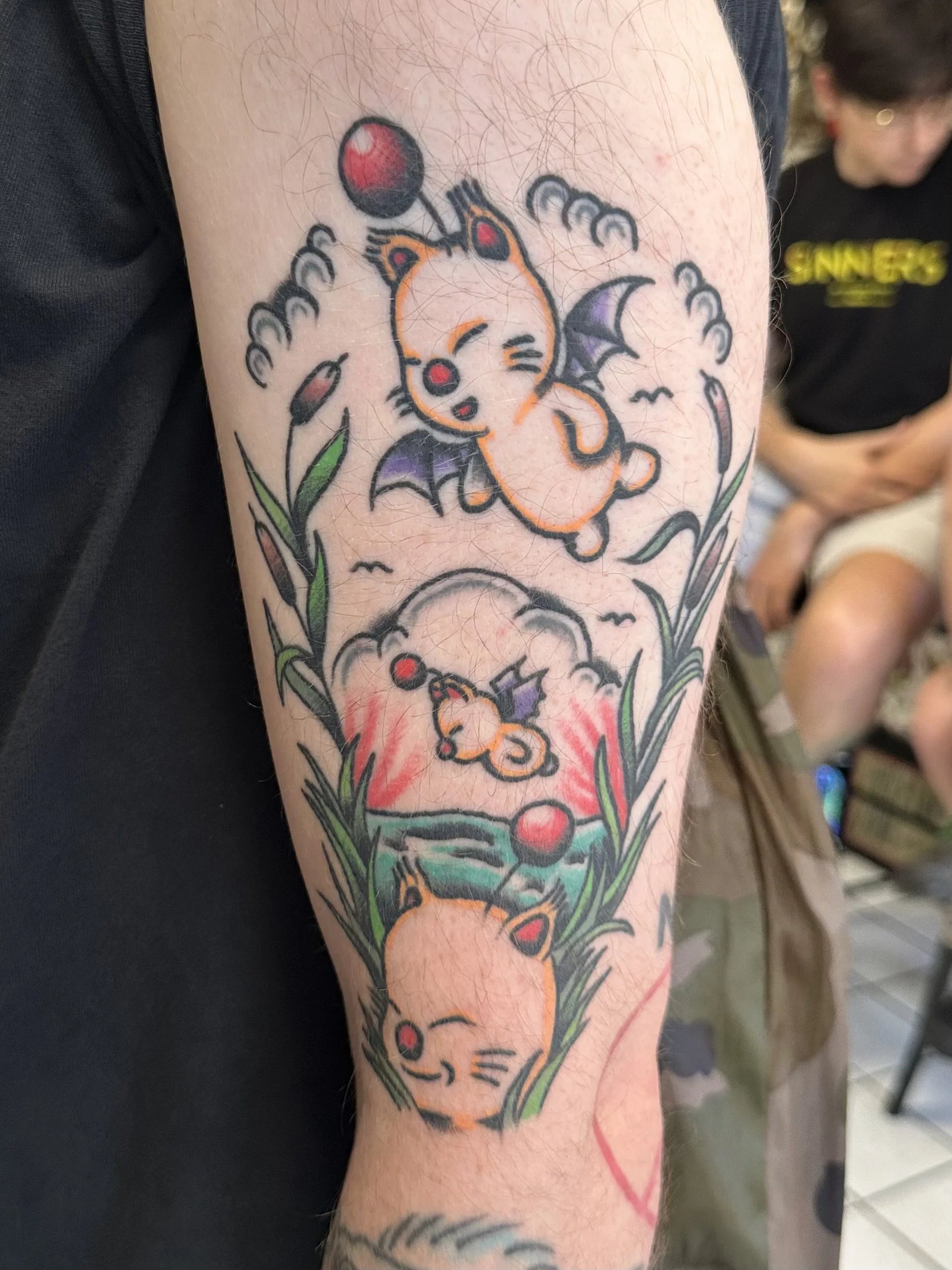 Colorful tattoo on arm featuring a cartoon-style orange kitten with bats wings, surrounded by grass, flowers, and red and black balloons.