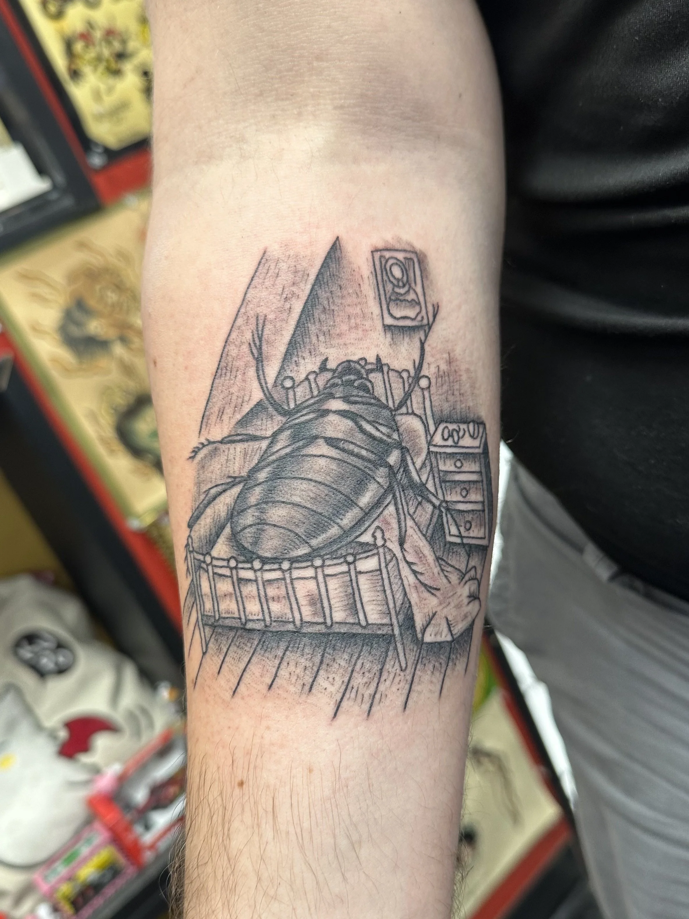 Black and gray tattoo of a giant beetle sitting on a bed in a room, with a framed picture on the wall behind it.