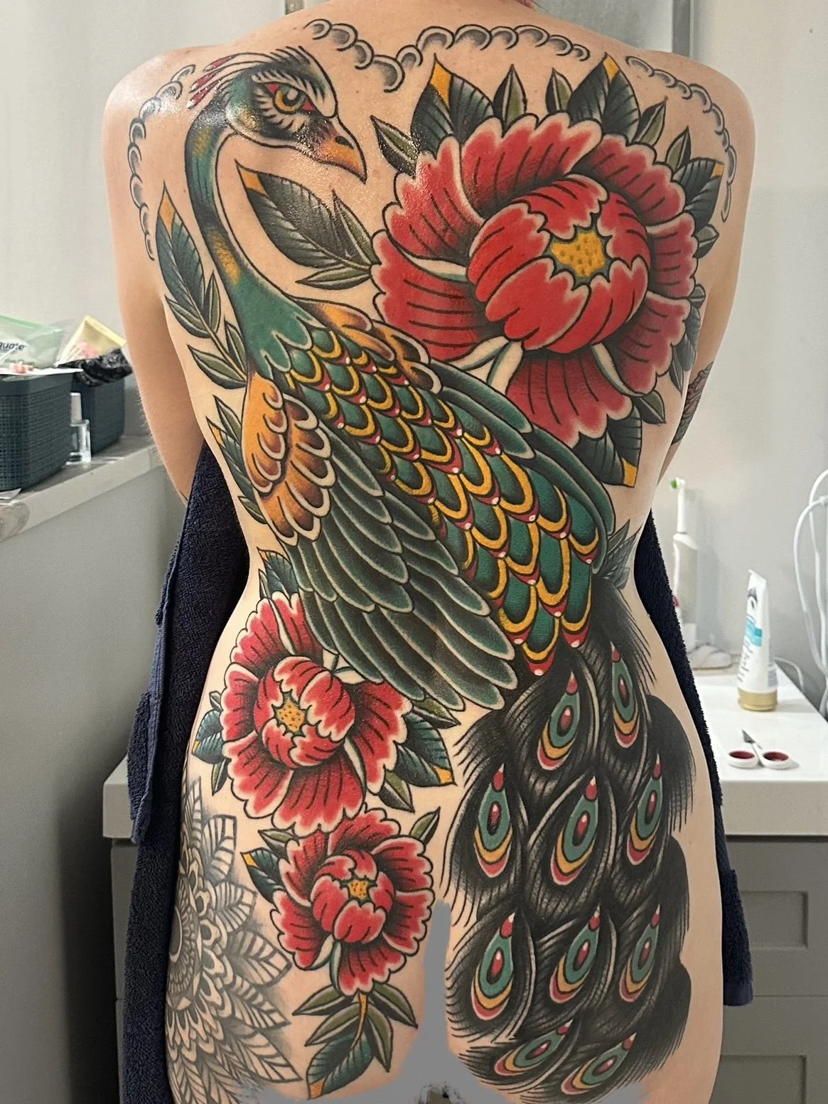 Colorful traditional-style tattoo of a peacock with vibrant feathers and large red flowers on the back of a person.