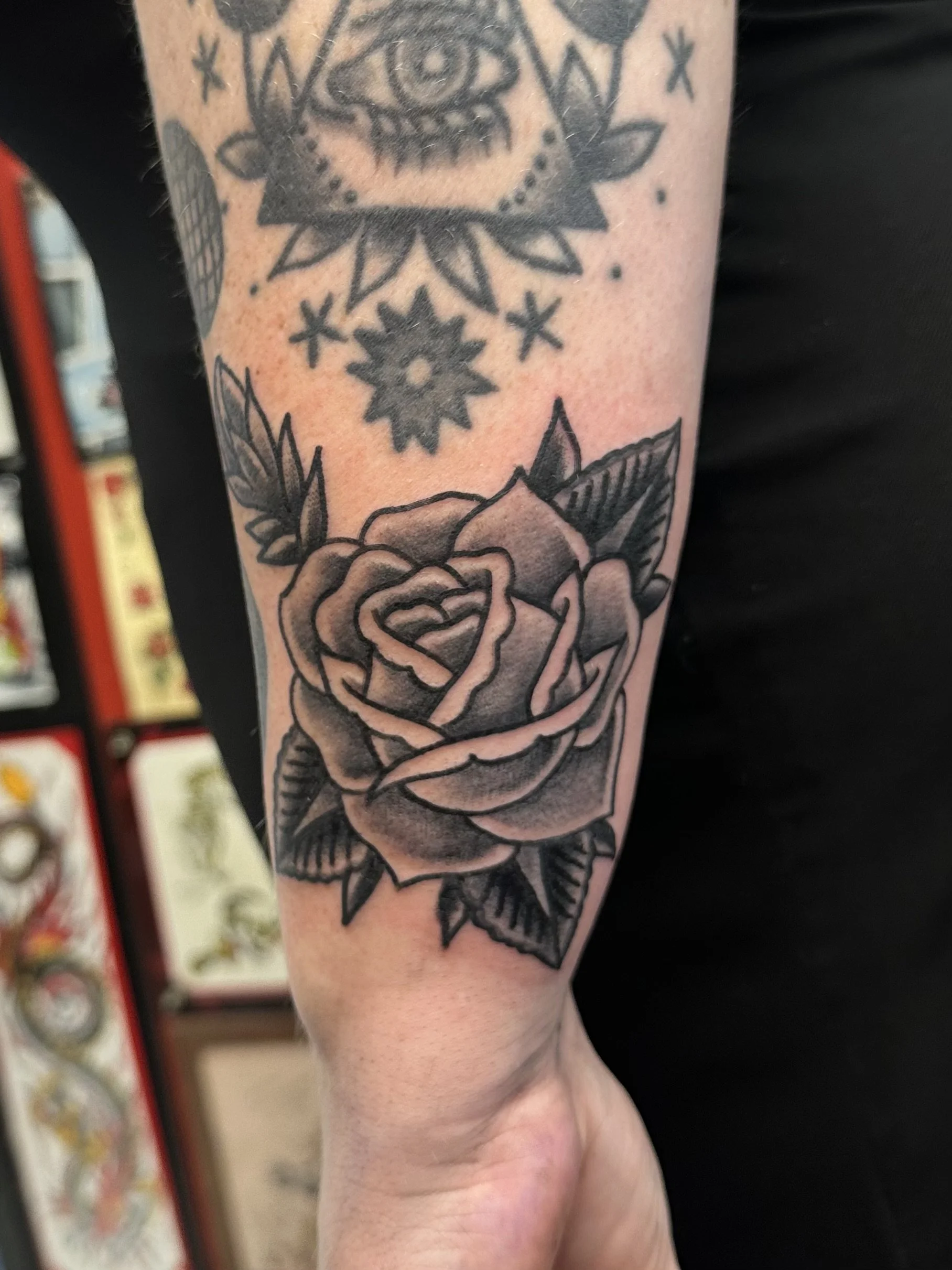 A black and gray tattoo on a person's arm featuring a rose with detailed petals and leaves at the bottom, a geometric star-like shape with multiple points, a small star, a sun symbol with a circle in the center, and an eye with a triangular design ar
