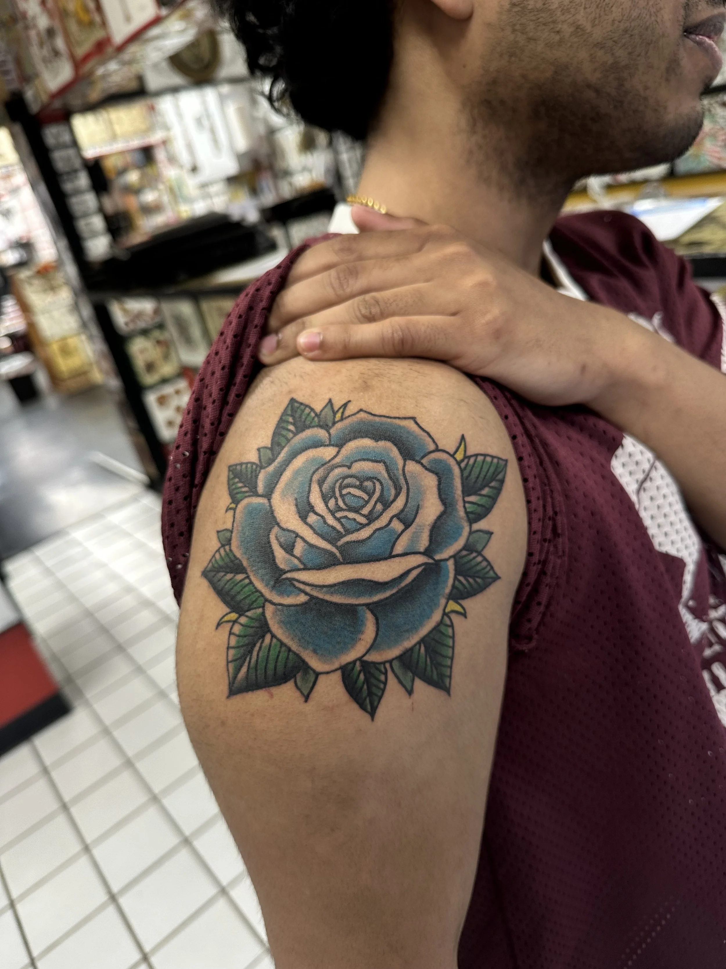 A person displaying a colorful tattoo of a blue and peach rose on their upper arm. The individual is wearing a maroon sleeveless shirt and a gold necklace. They are holding their shoulder with one hand, and the photo is taken in a store with shelves 