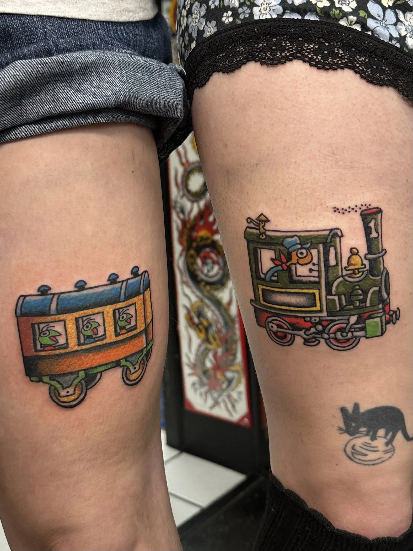 Close-up of two people's thighs showing colorful tattoo designs of toy trains on their skin.