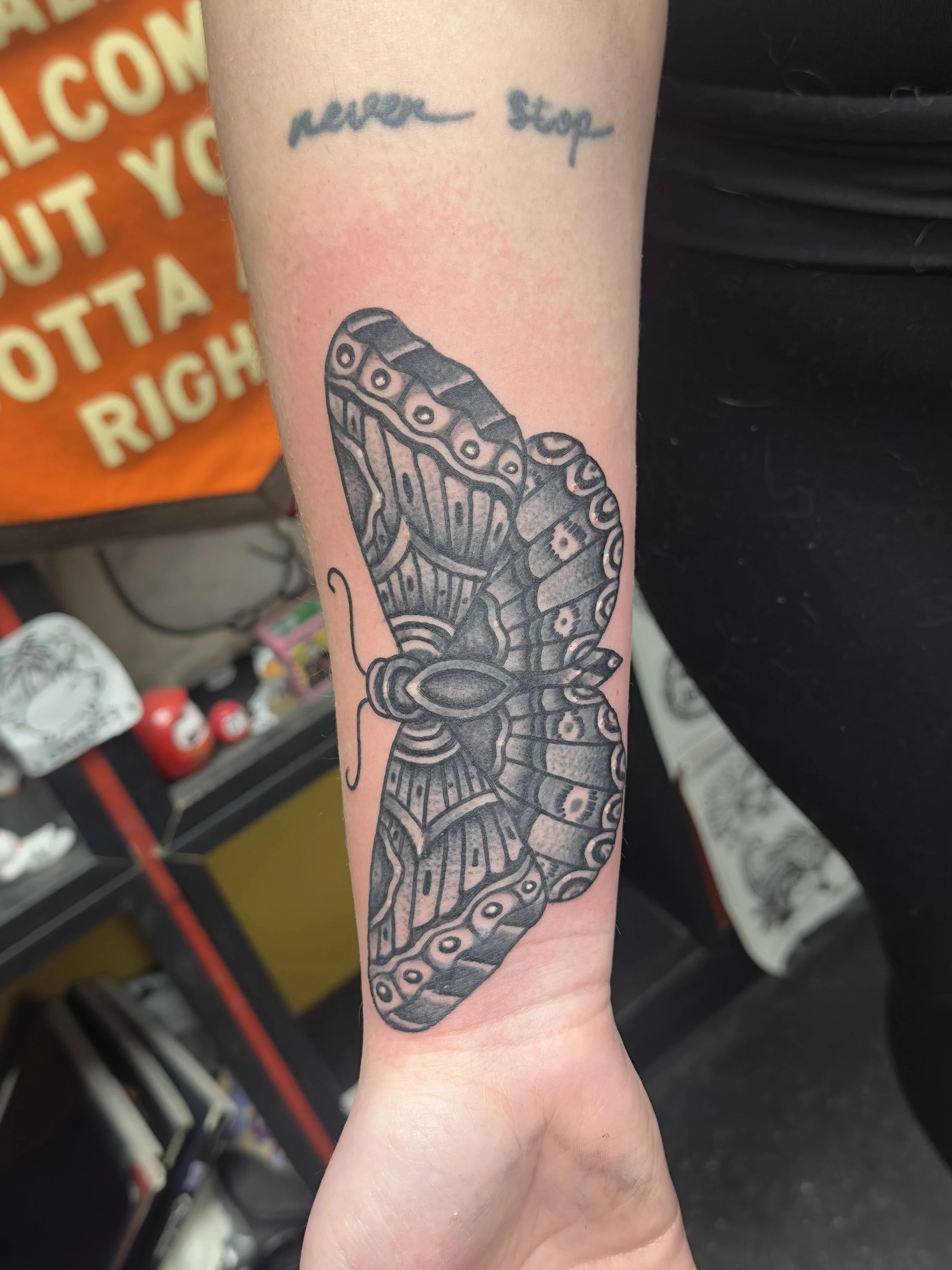 Black and gray tattoo of a butterfly on person's arm, with text "never stop" in cursive above it.