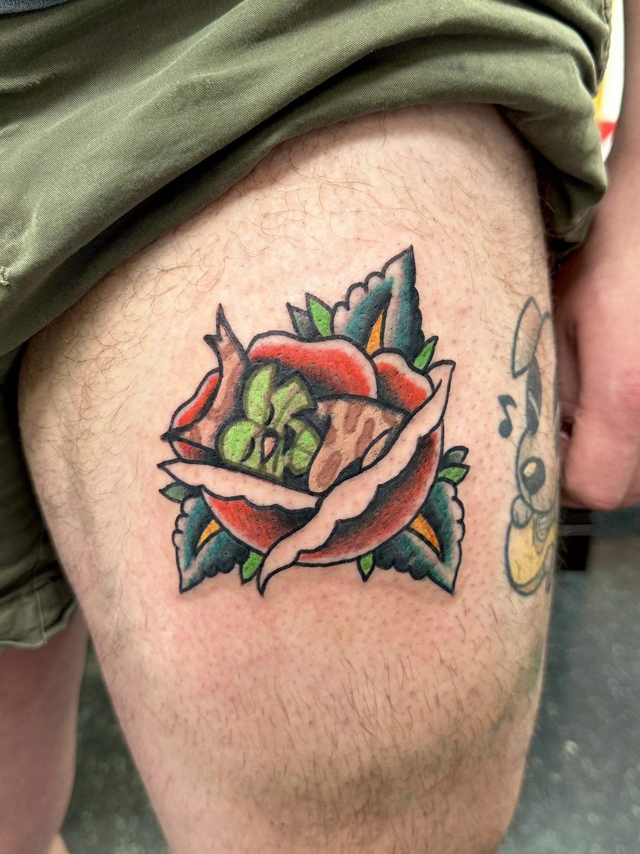 A tattoo of a colorful rose with green leaves on a person's upper thigh, with part of the person's green shorts visible at the top of the image.