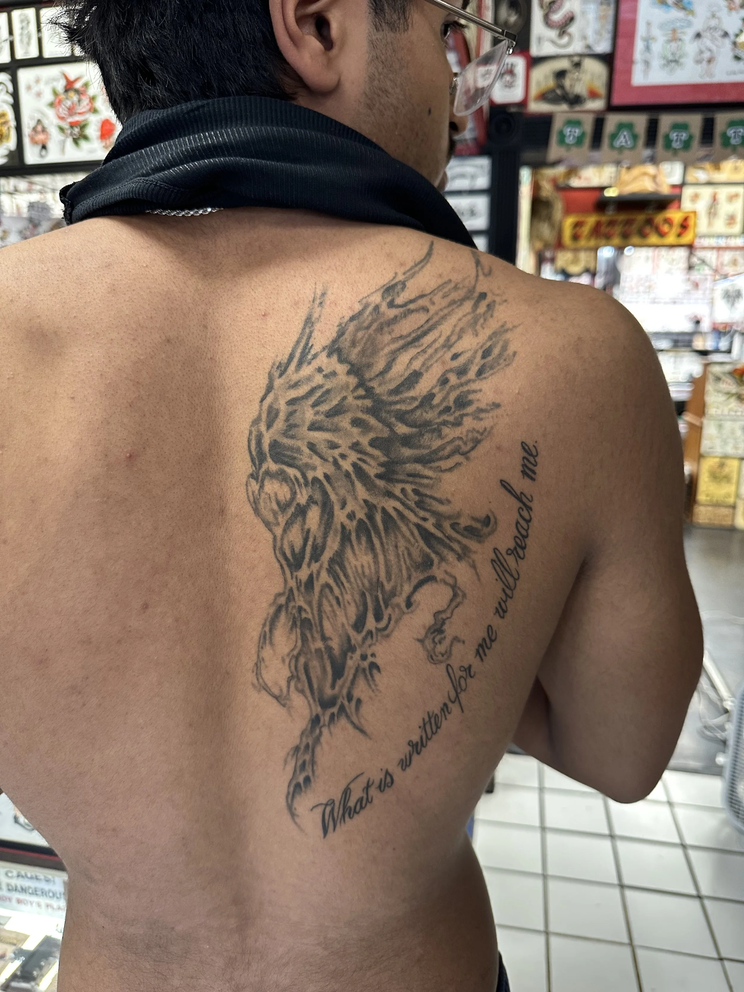 A person with glasses and short dark hair showing a large tattoo of a lion's head on their shoulder and upper chest, with a quote beneath it, in a tattoo parlor with tattoo designs on the wall.