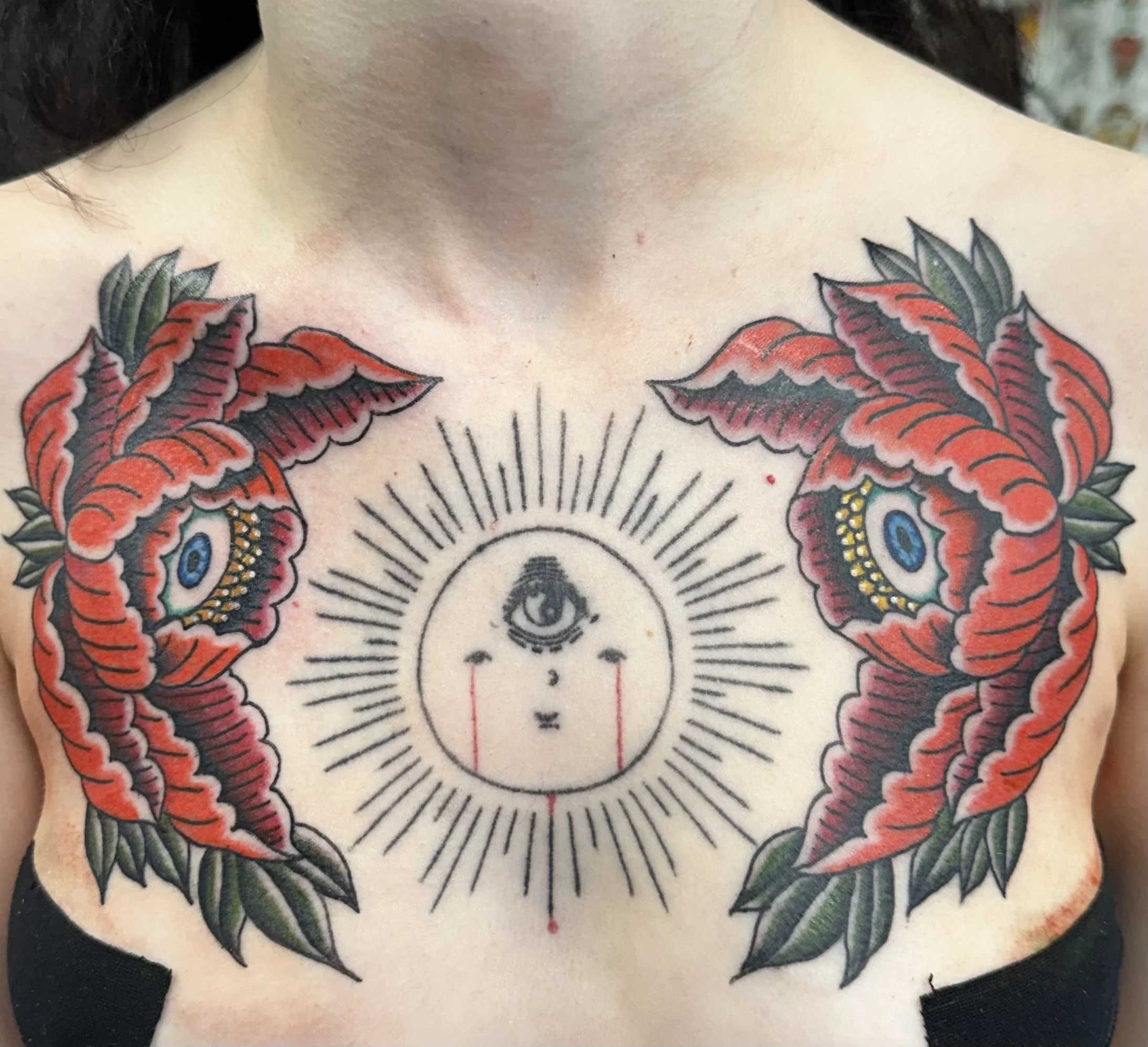 Close-up of a tattoo on a person's chest featuring a detailed sun with a realistic eye in the center, surrounded by colorful leaves and floral designs.