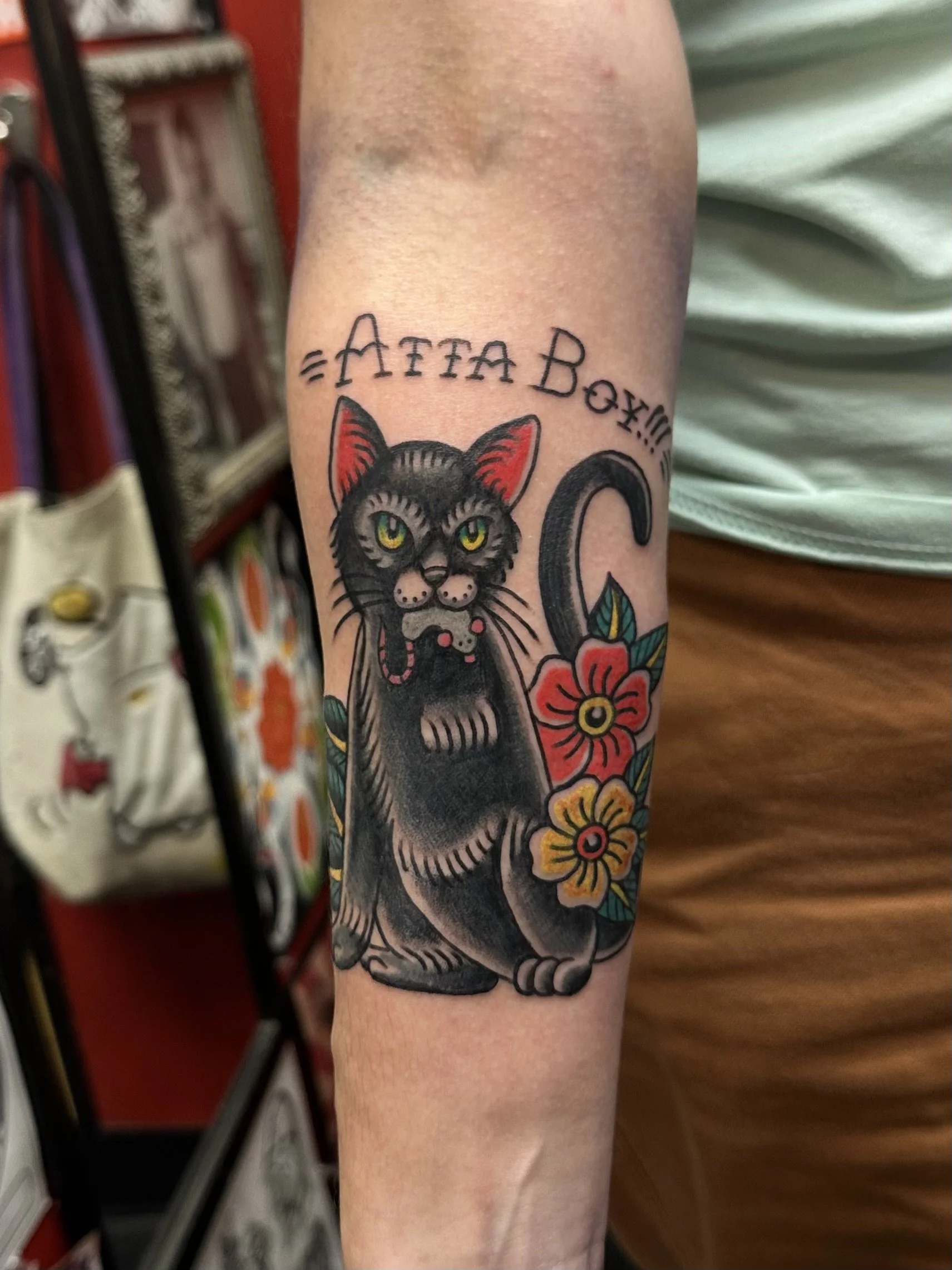 A tattoo of a black cat with yellow eyes surrounded by colorful flowers on a person's arm, with the phrase '=ATTACK!!=' written above in a stylized font.
