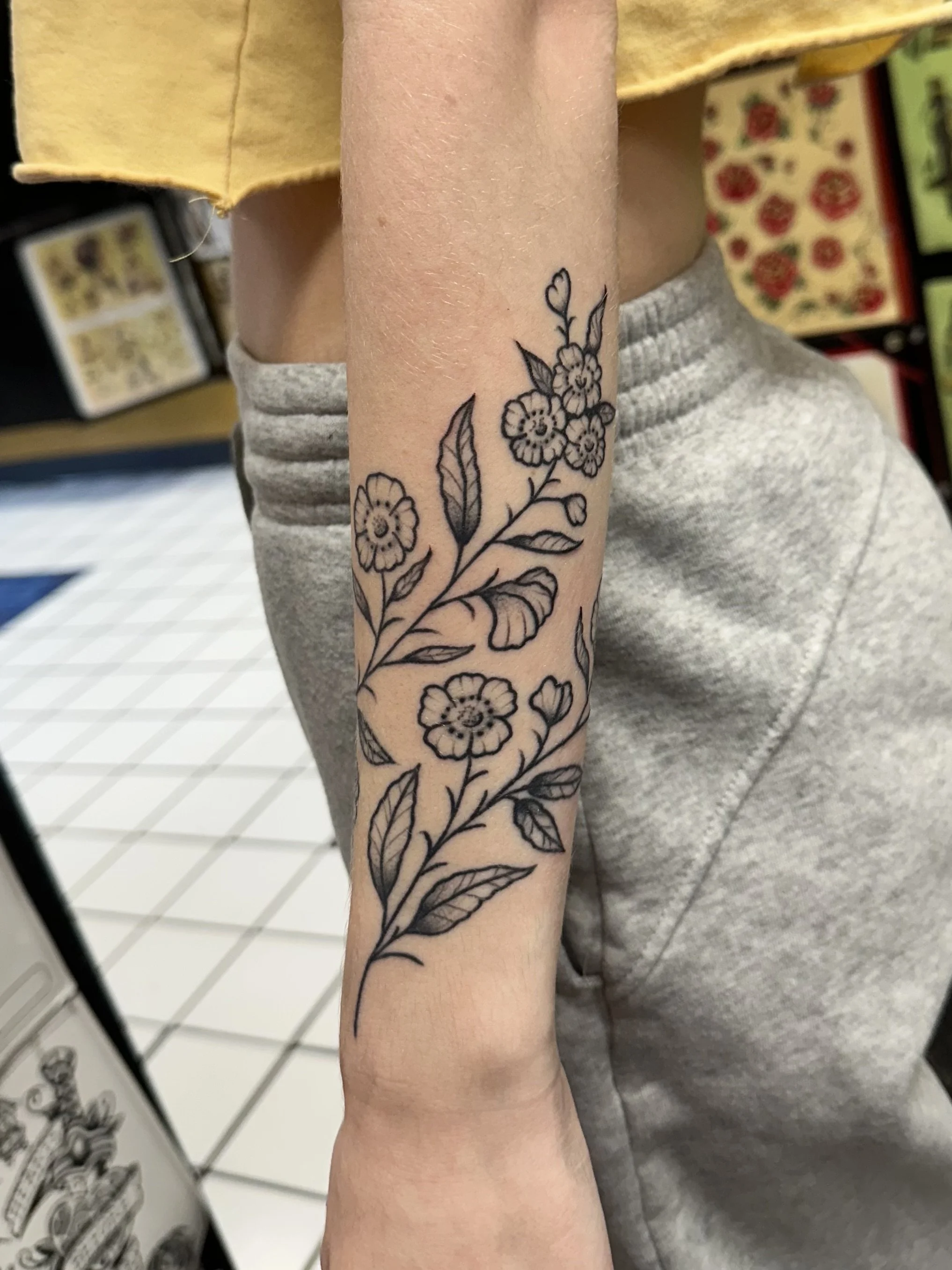 A black outline tattoo of a floral branch with flowers and leaves on a person's forearm.