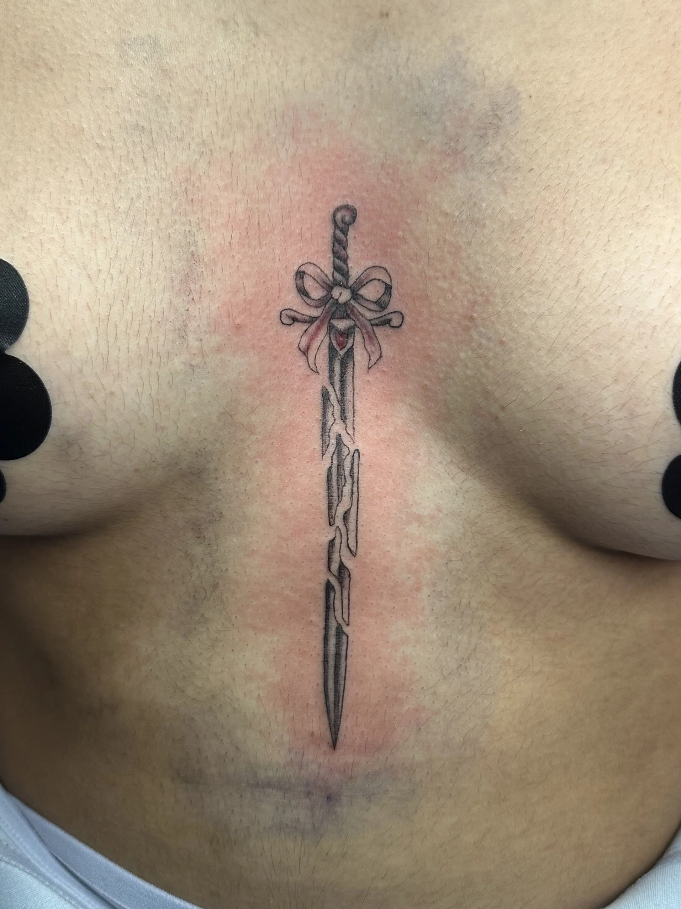 Tattoo of a sword with a bow tied around the hilt, located in the center of the person's chest.