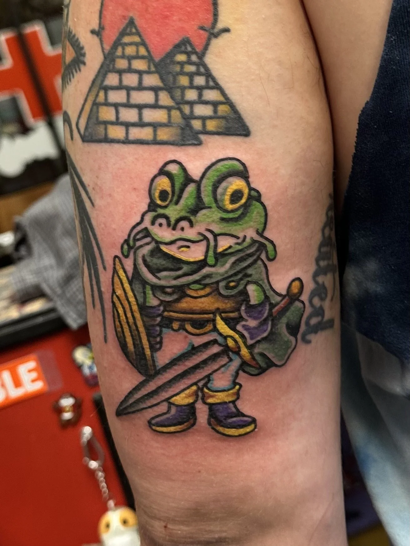 Tattoo of a cartoon frog dressed as a knight, holding a sword and shield, with geometric shapes resembling pyramids and a sun in the background.