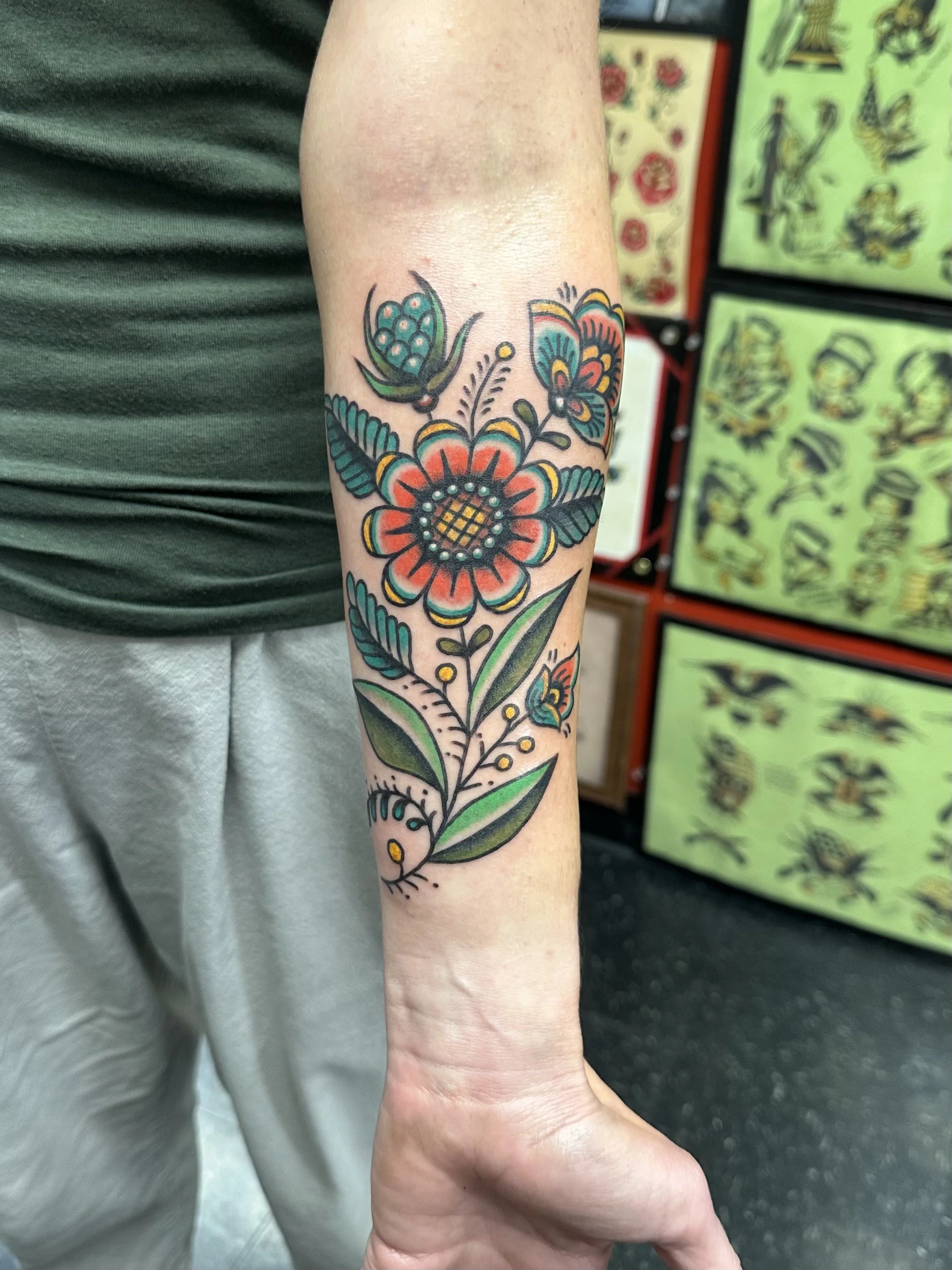A colorful floral tattoo on a person's forearm, featuring a large orange and yellow flower with black outlines, surrounded by green leaves and smaller decorative elements, against a background of tattoo designs in a shop.