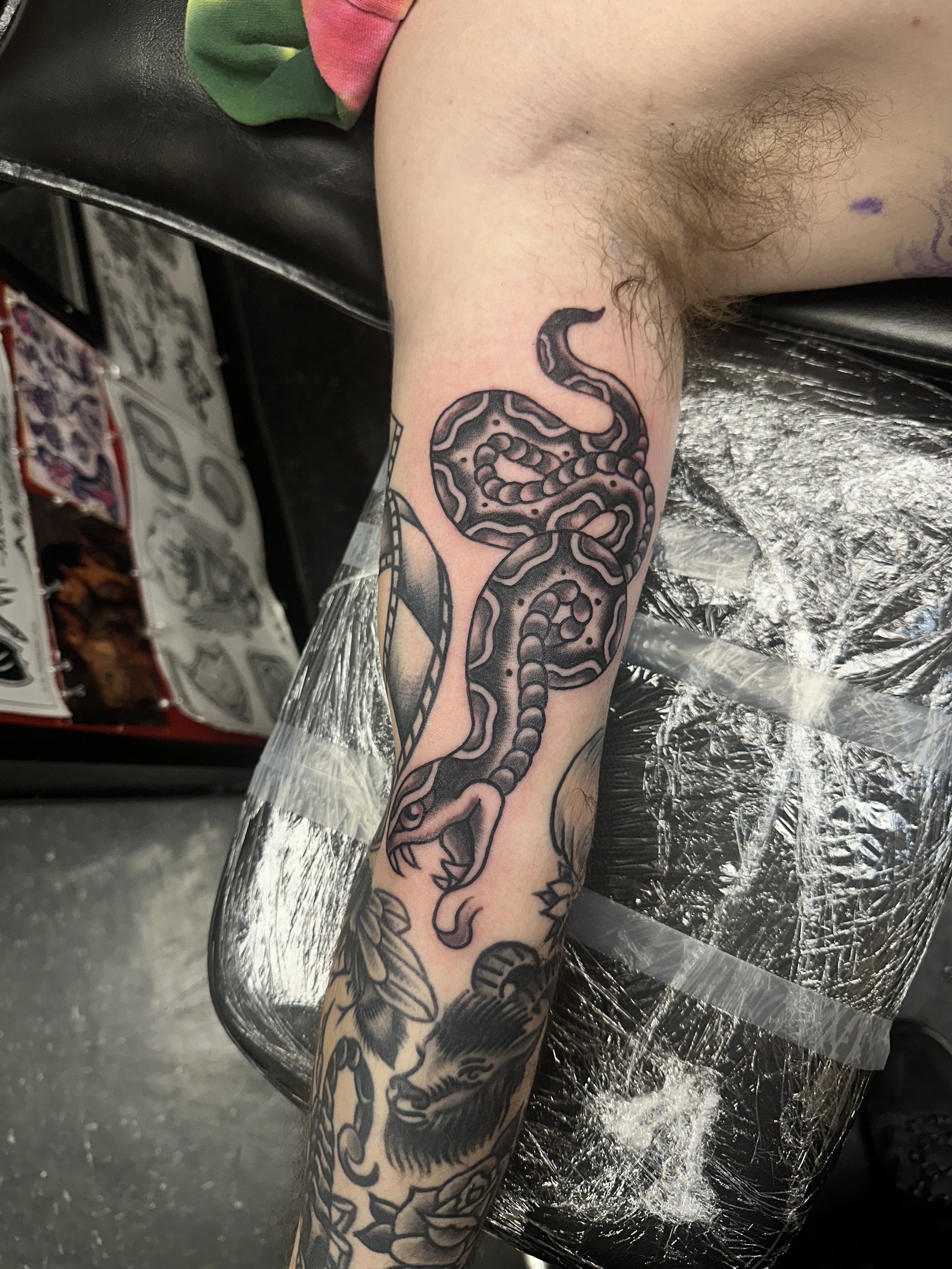 A tattoo of a snake on a person's arm, with additional body art visible, seated on a black chair with plastic coverings, at a tattoo studio.